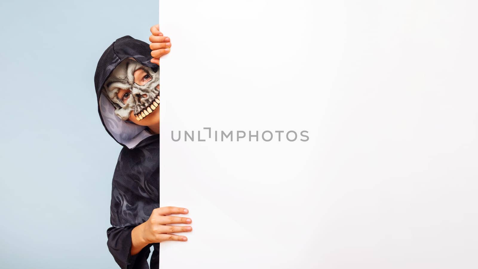 Happy Halloween. Cute little boy in a costume hiding behind white banner blank. Happy kid holding cardboard background with copy space.