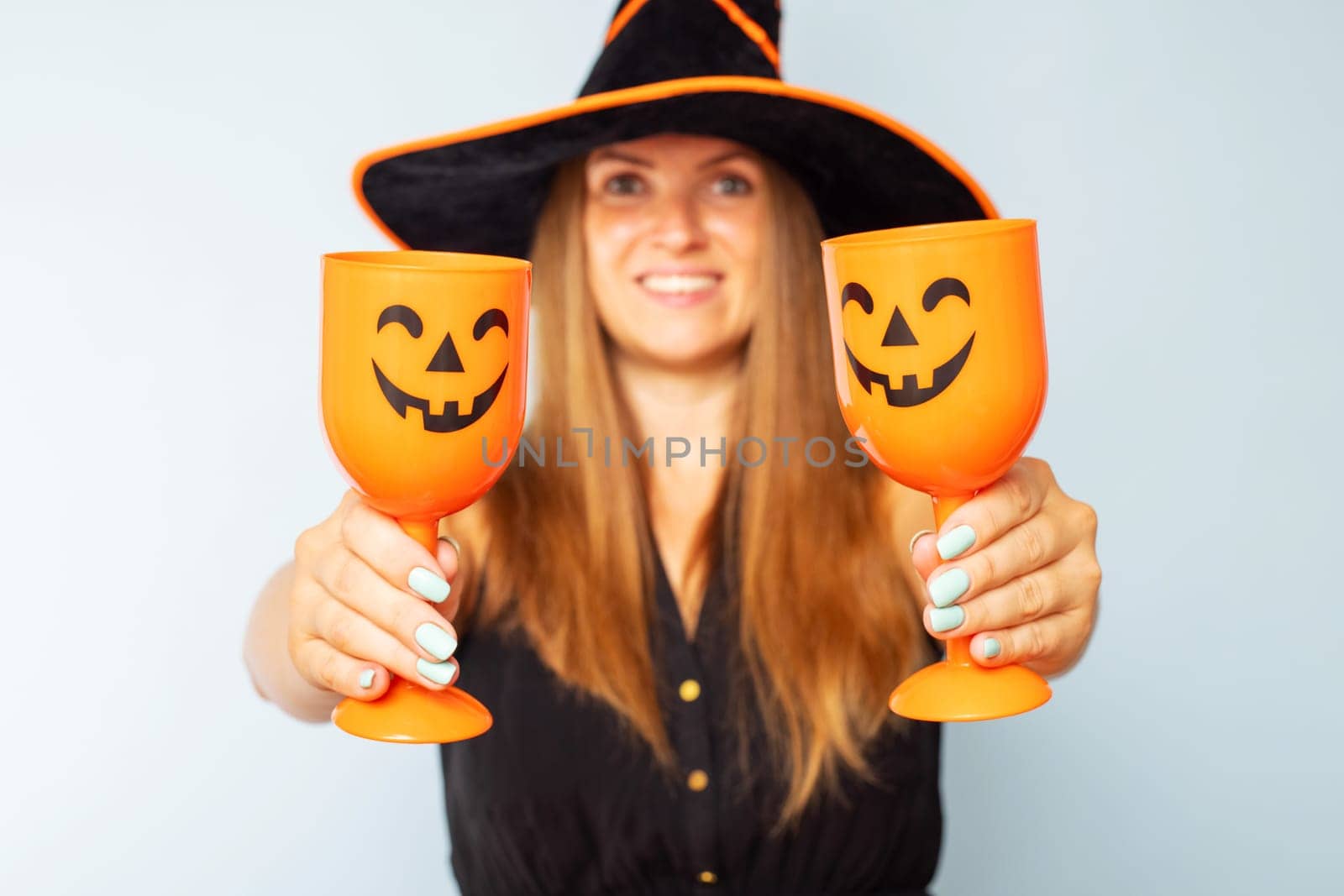 Happy Halloween. Happy brunette woman in halloween witch costume with black hat holding glasses of cocktails on a light background. Halloween party.