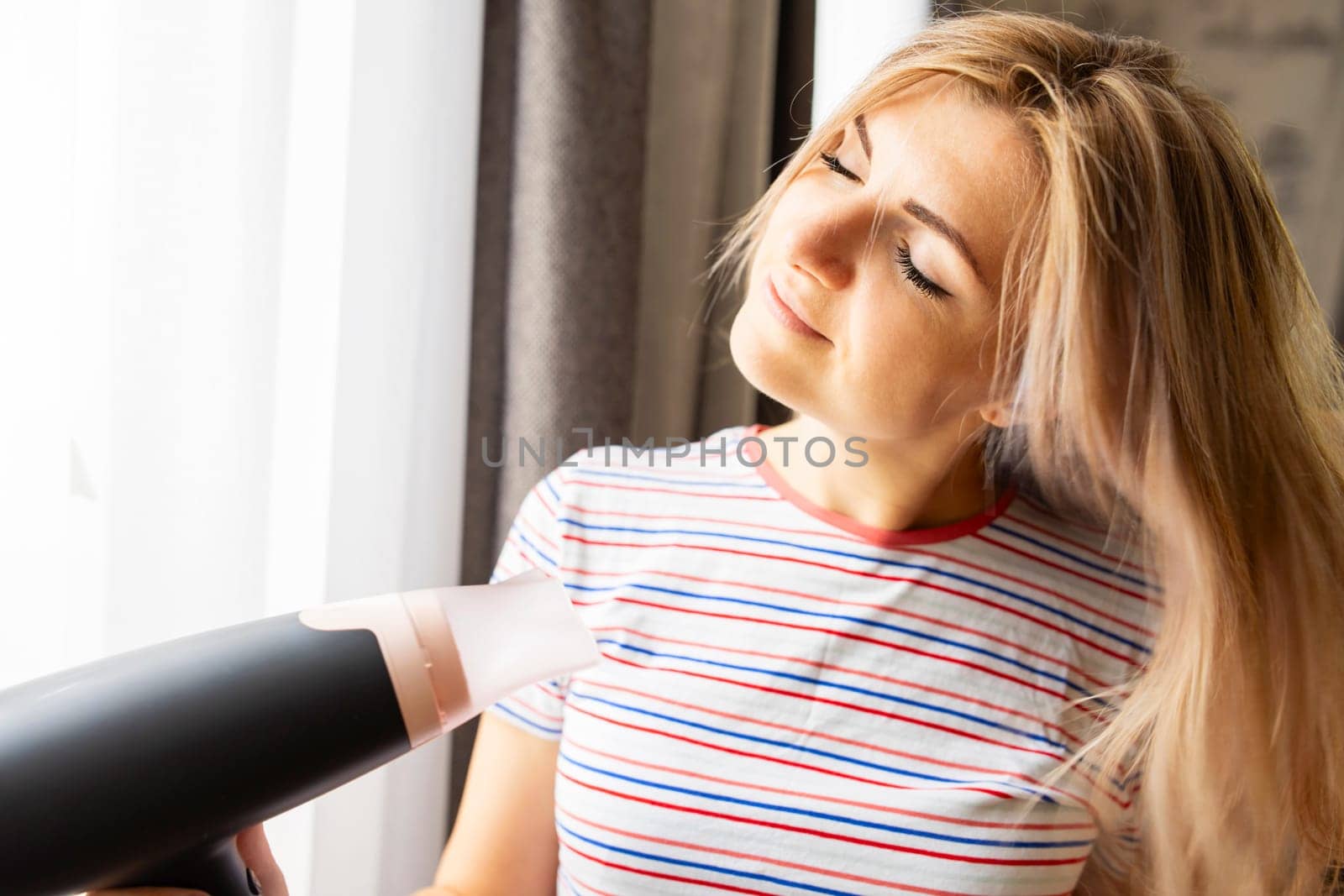 Beautiful young woman using hair dryer near the window at home.