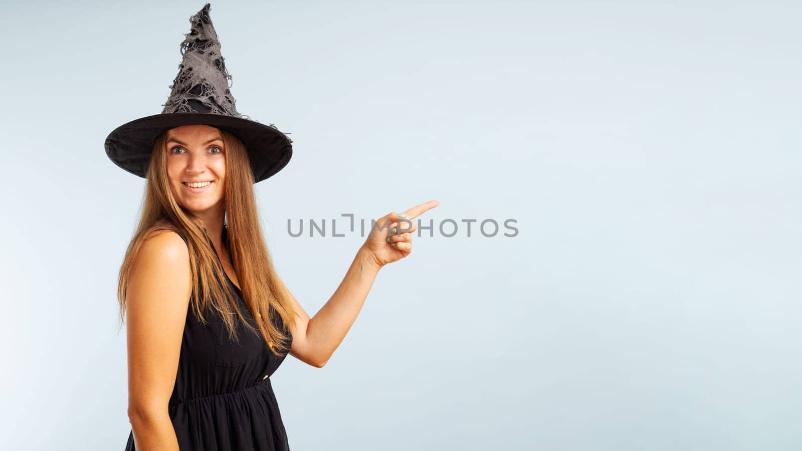 Woman in witch costume pointing with finger at copyspace side and looking at camera on blue background. Happy Halloween.