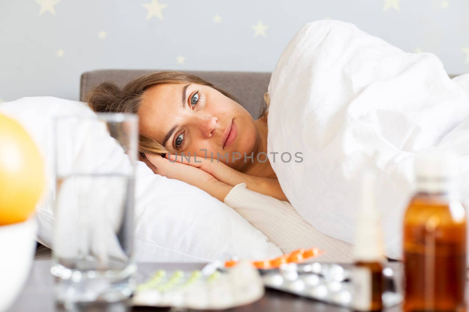 Sick young woman covered with a blanket lying in bed with high fever and a flu. A lot of medicines on table in bedroom.