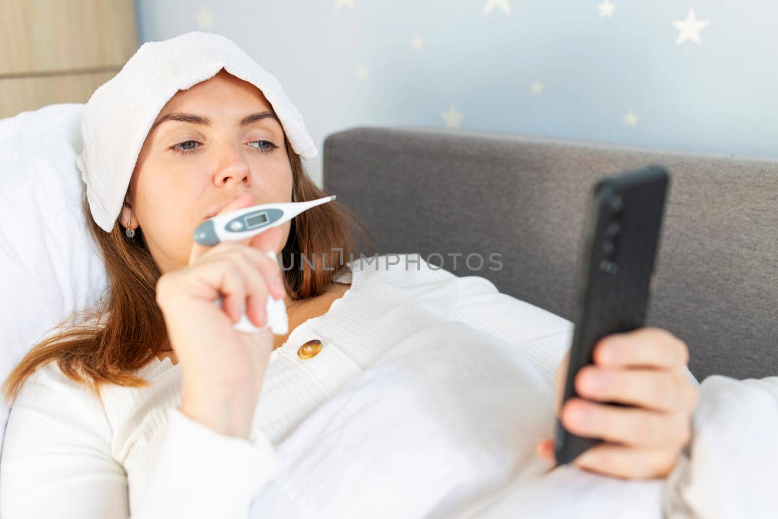 Sick young woman with bad cold, flu or coronavirus fever holding thermometer and talking to telemedicine service doctor. Sick woman having online medical consultation video call.