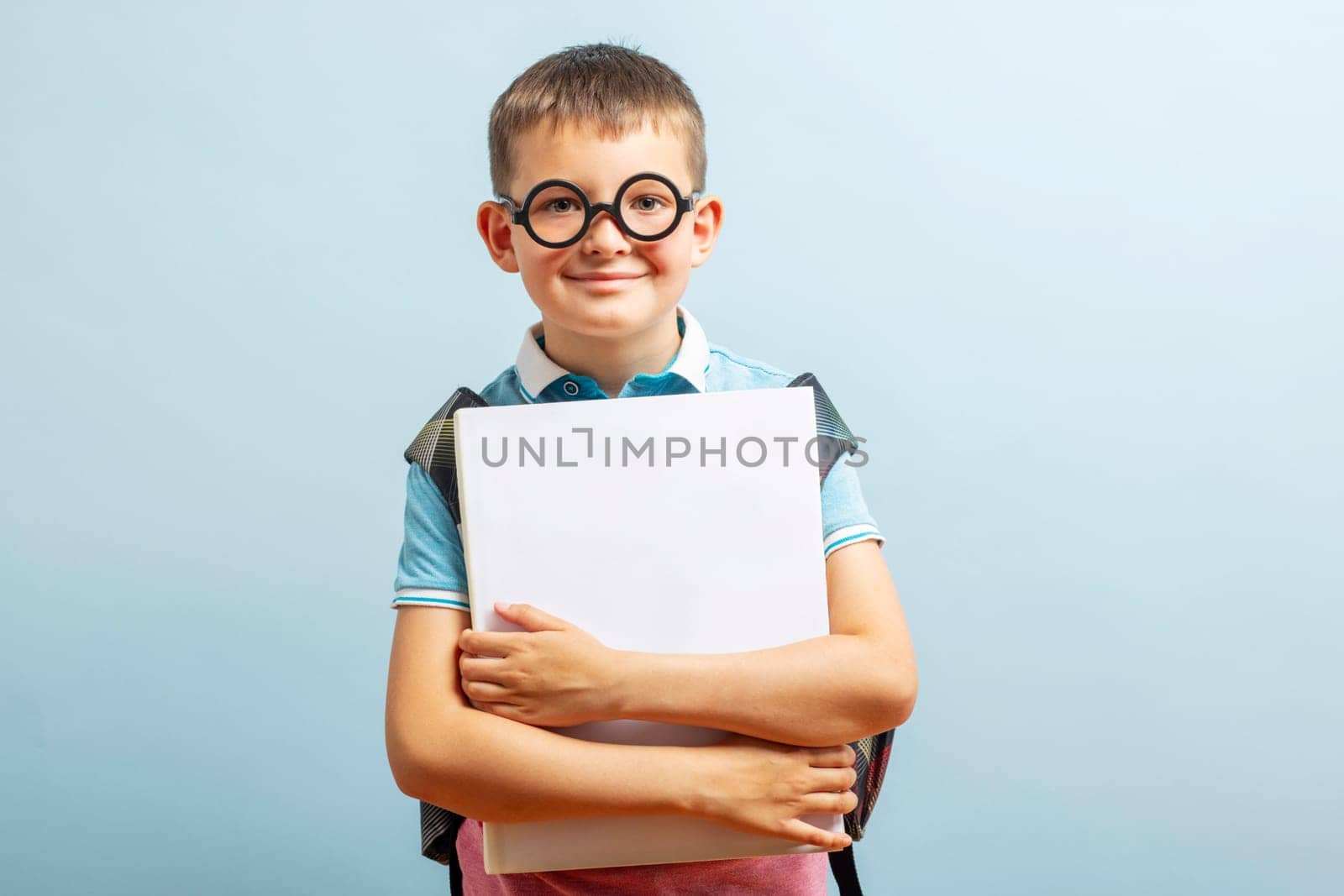 Back to school. Happy elementary school student in eyeglasses hugging a book smiling at camera on blue background. Free space.
