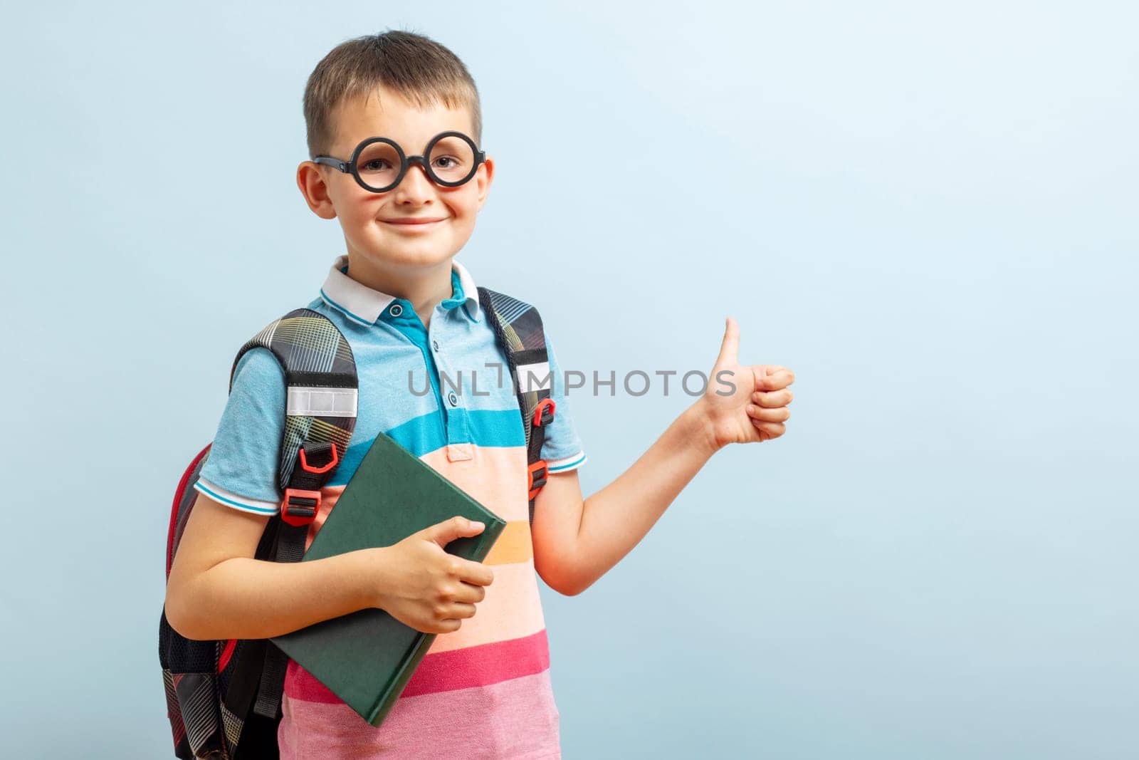 Back to school. Happy elementary school student in eyeglasses hugging book and showing a thumbs up gesture on blue background. Free space.
