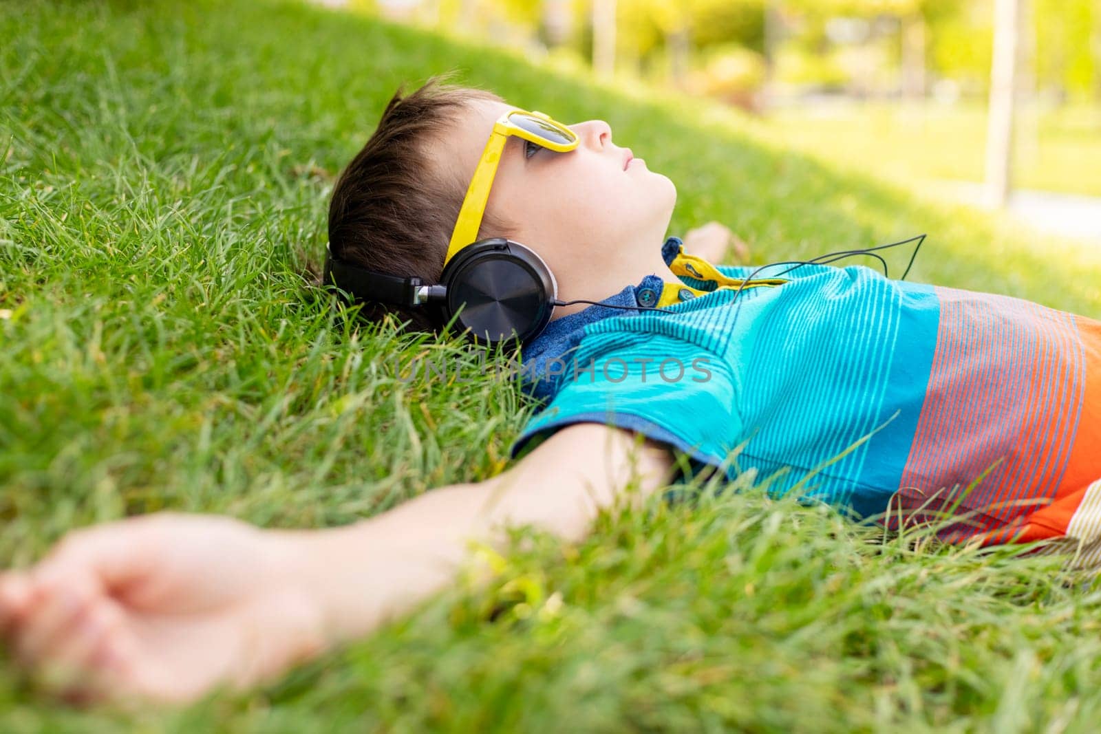Happy boy in sunglasses lying on grass and listening to music in headphones by andreyz