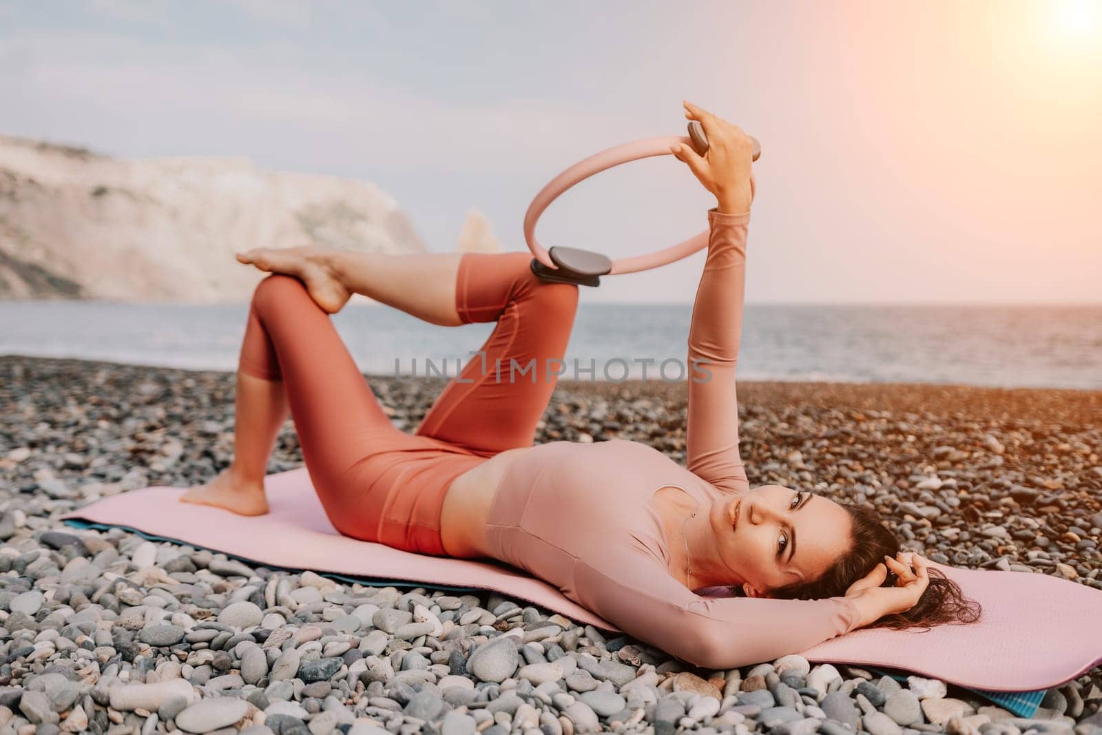 Middle aged well looking woman with black hair doing Pilates with the ring on the yoga mat near the sea on the pebble beach. Female fitness yoga concept. Healthy lifestyle, harmony and meditation.