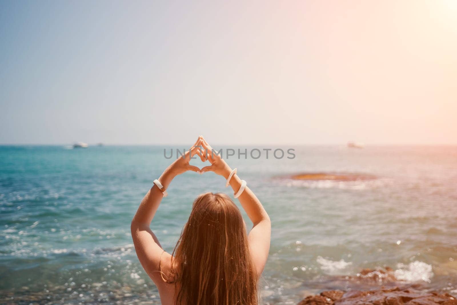 Woman makes heart with hands on beach. Young woman with long hair in white swimsuit and boho style braclets practicing by sea ocean. Concept of longing daydreaming love peace contemplation vacations by panophotograph