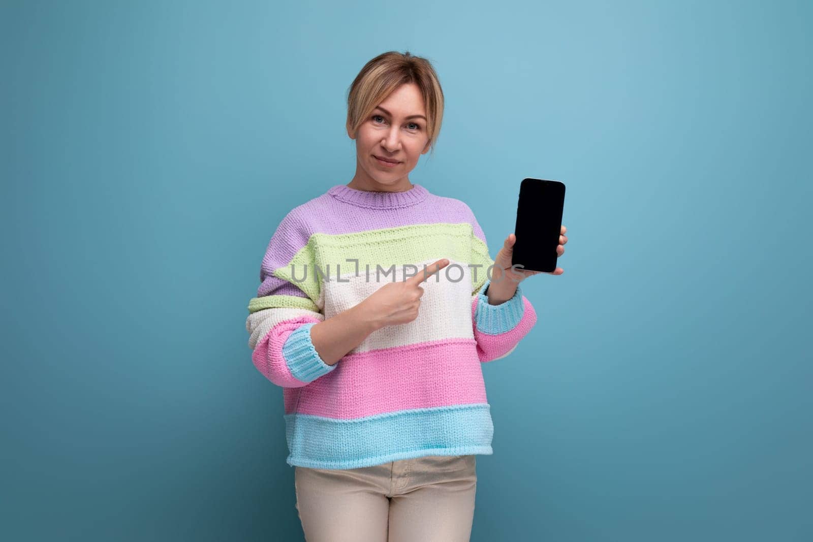 pleasant blond young woman in a striped sweater demonstrates a smartphone mockup on a blue background.