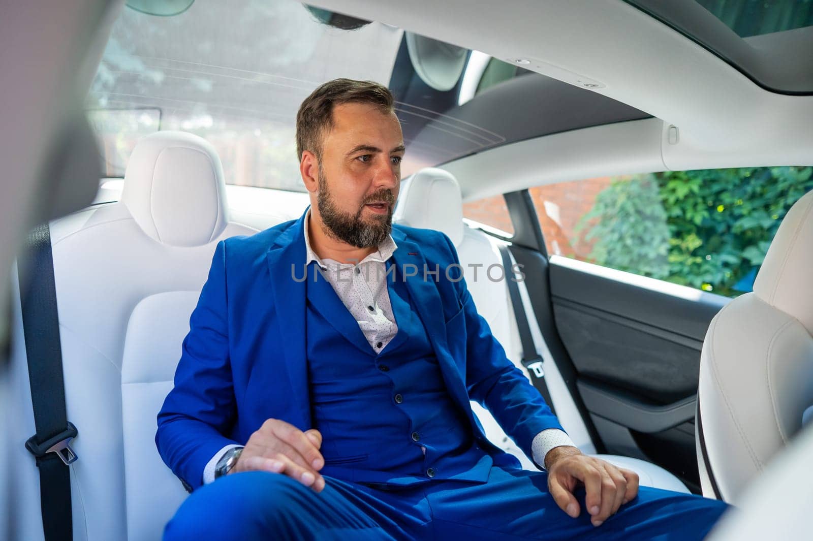 A caucasian man in a blue suit sits in the back seat of a car. Business class passenger