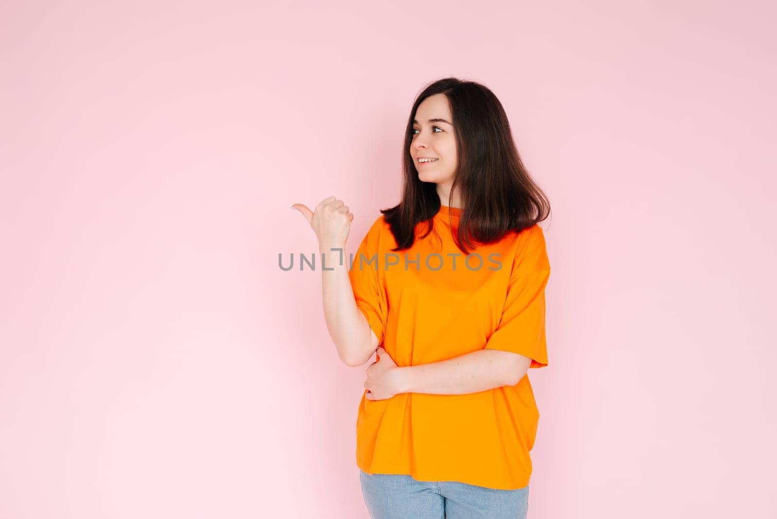 Alluring Advertising Space: Charming Lady Pointing Thumbs at Empty Area - Attention-Grabbing Concept for Promotional Opportunities on Pink Background