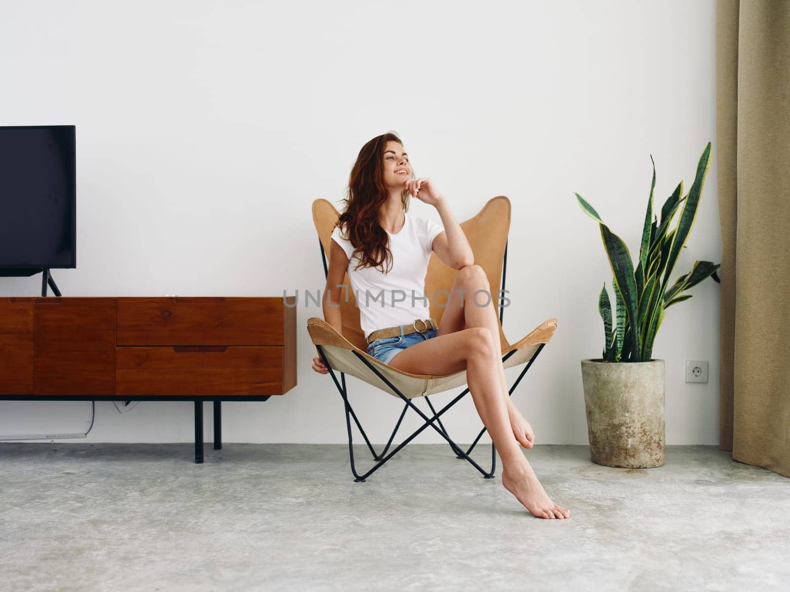 Woman sitting in leather armchair smile with teeth Lifestyle in white T-shirt and blue denim shorts, resting at home stylish modern interior with white walls, copy space. by SHOTPRIME