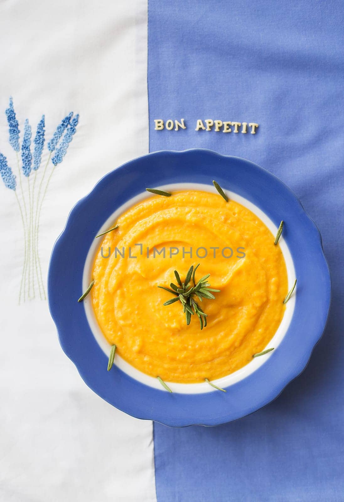 pumpkin soup in a blue plate with rosemary on a napkin with flowers, lettering bon appetit