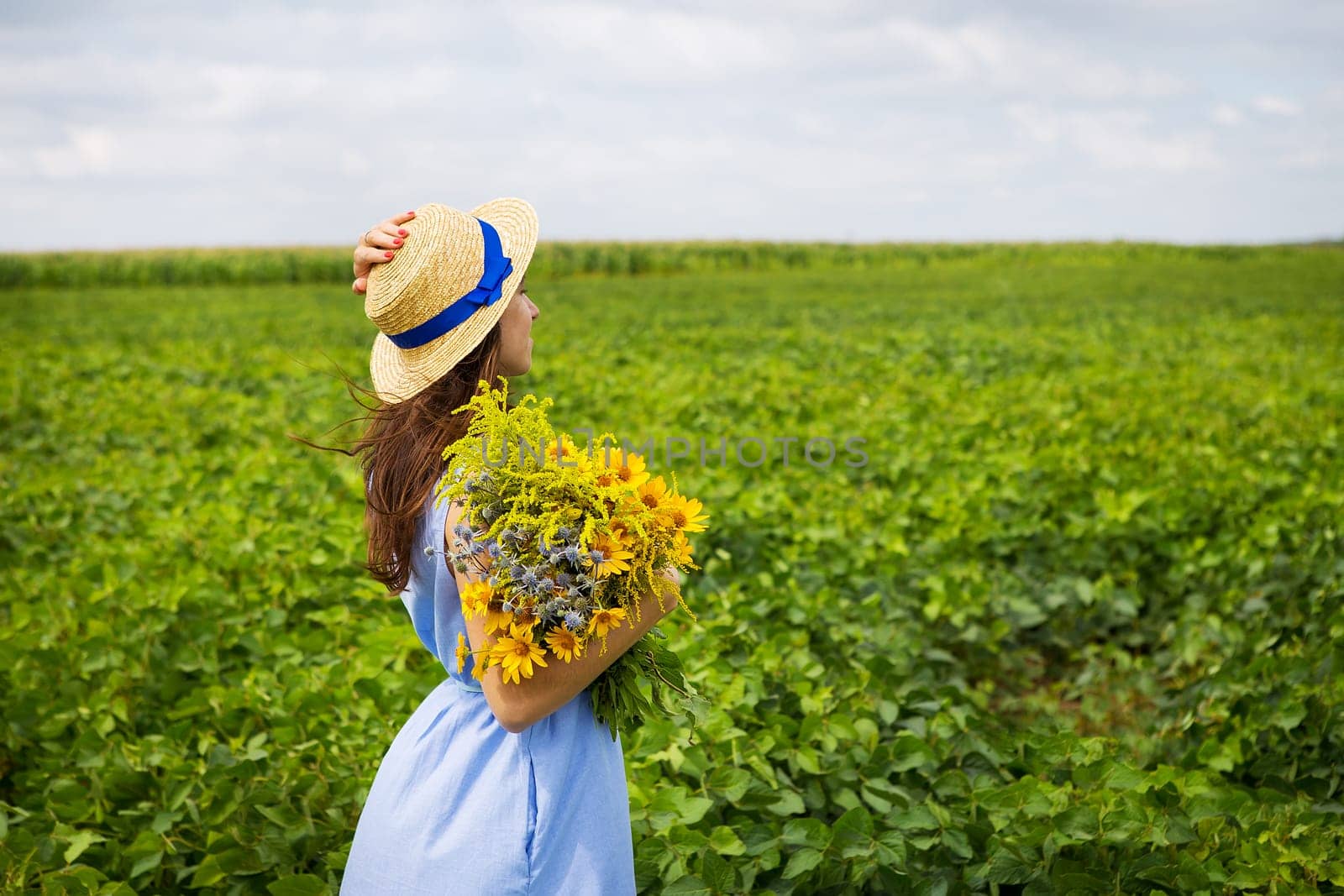 beautiful girl stands in the field with a bouquet of yellow flowers and a straw hat.