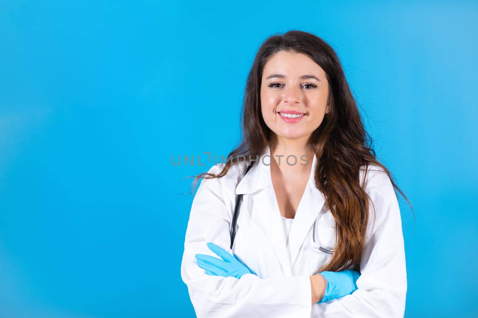 Long-haired woman crosses arms with smiling and delighted expression waiting for patient at blue wall. Young doctor with stethoscope enjoys working in health sphere