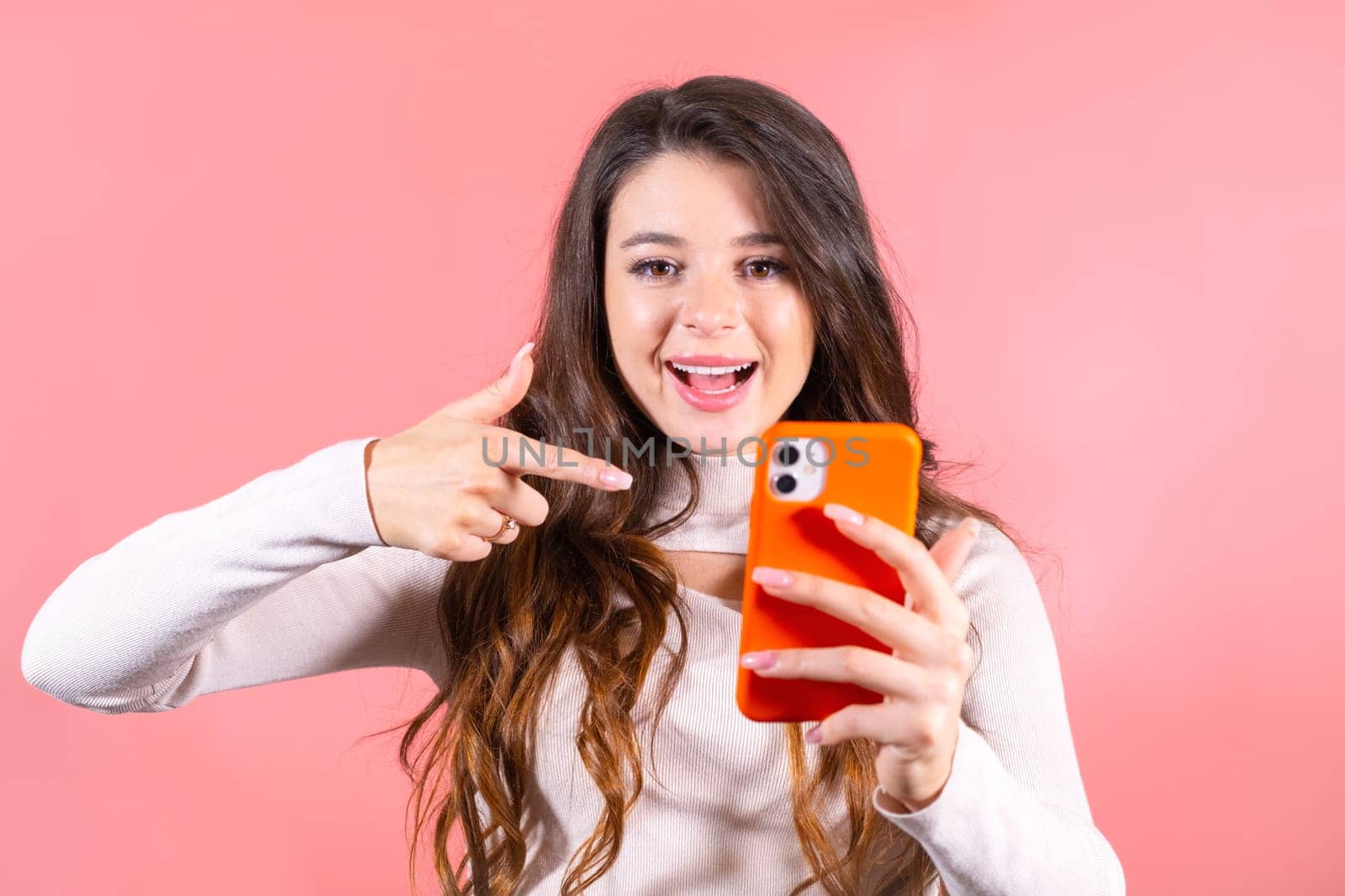 Young woman with delighted expression looks at smartphone pointing finger to wall. Brown-haired lady with amused expression at pink studio wall