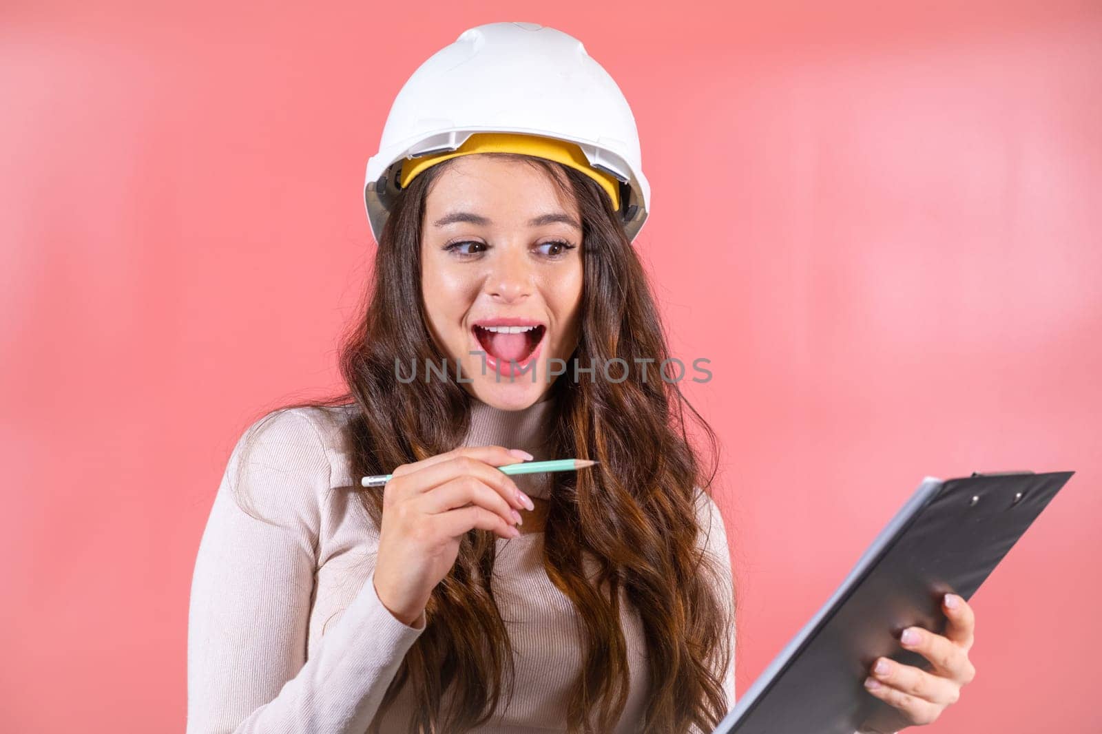 Woman wearing white hardhat writes information for building project on clipboard. Brown-haired architect with smiling and delighted expression enjoys job