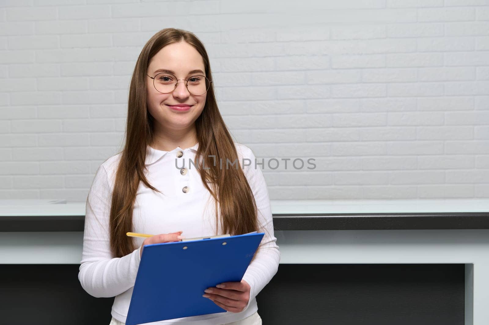 Confident portrait of Caucasian young pretty woman, purposeful receptionist administrator with clipboard near reception desk in modern clinic, smiling looking at camera, keeping a doctor's appointment