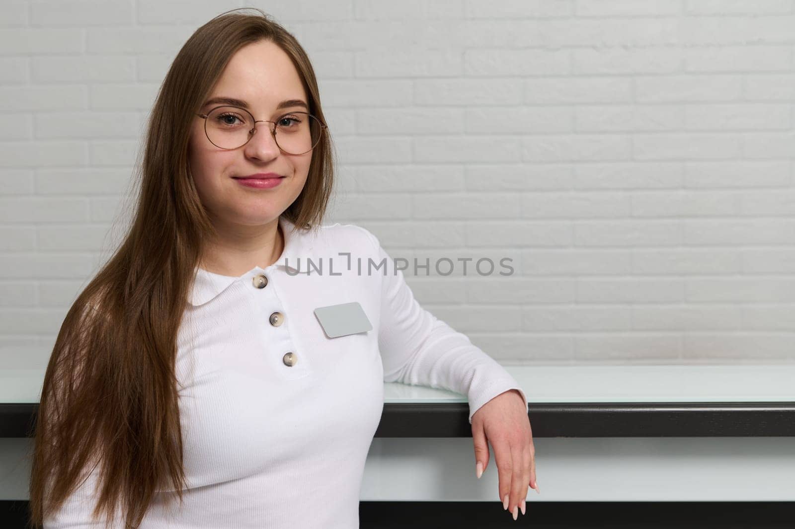 Smiling young receptionist in medical uniform with pinned blank badge, standing at the reception desk in modern medical clinic, outpatient hospital, dentistry office of beauty spa center. Copy space