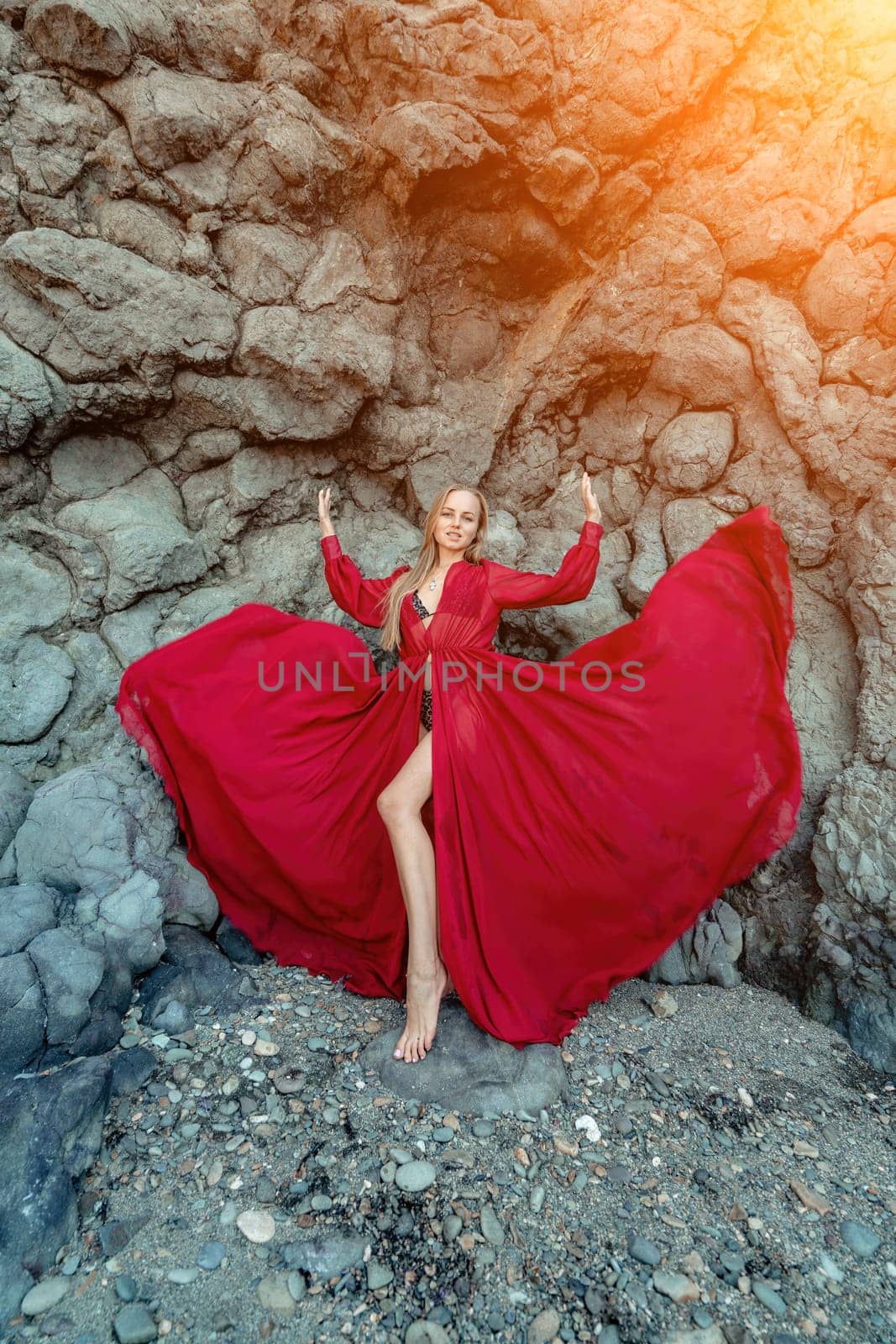 Red dress rocks woman. A blonde with flowing hair in a long flowing red dress stands near a rock of volcanic origin. Travel concept, photo session at sea by Matiunina