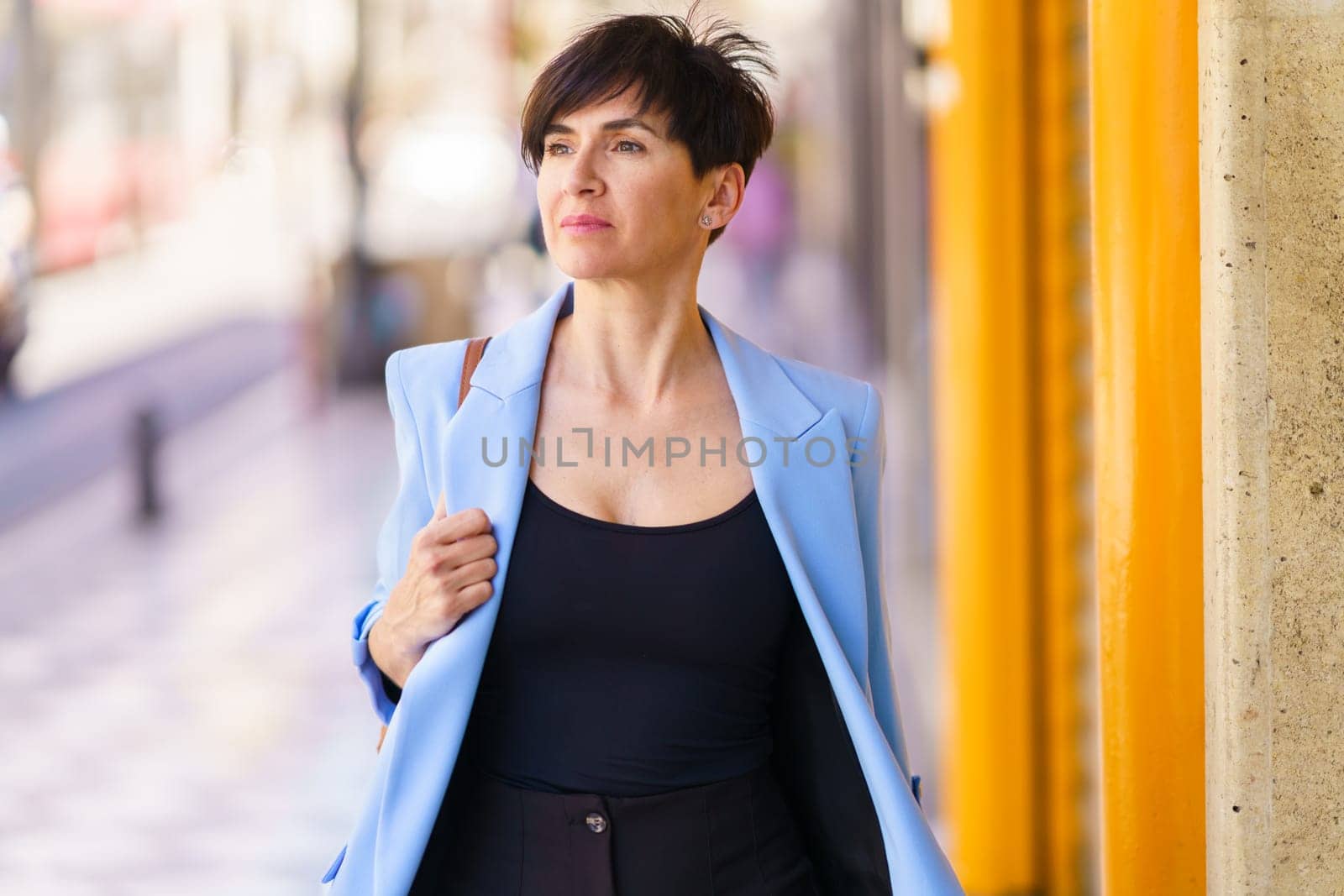 Portrait of confident female entrepreneur in formal clothes with handbag standing on blurred background street near yellow entrance and looking away