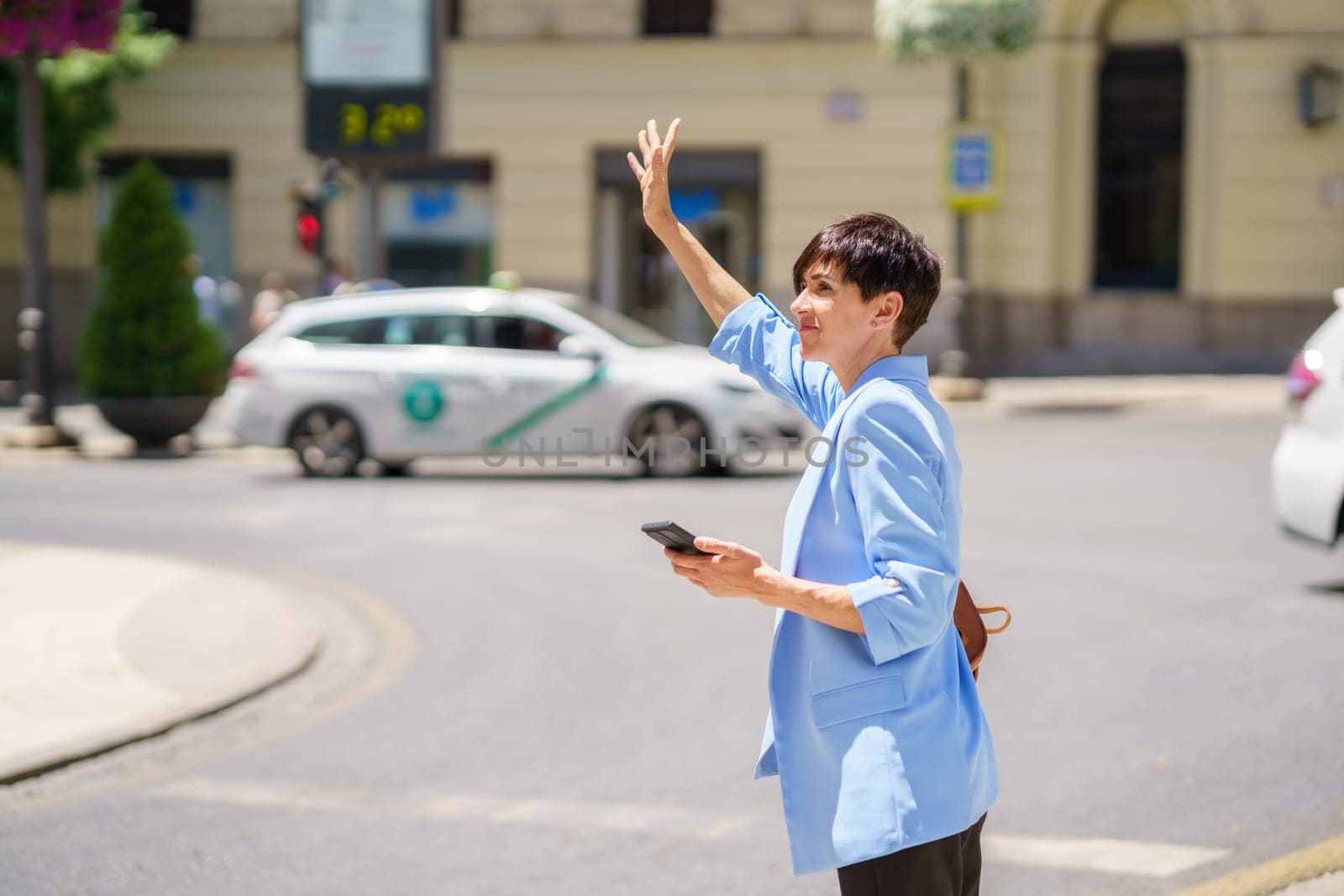 Side view of serious female in formal clothes standing on street junction and waving hand to stop taxi while holding mobile phone in bright daylight