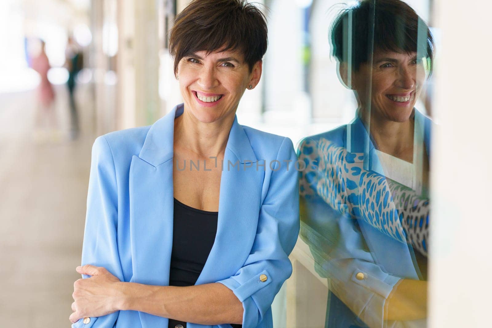 Positive middle aged female in blue jacket with short brown hair looking at camera and smiling while leaning on glass wall of modern building