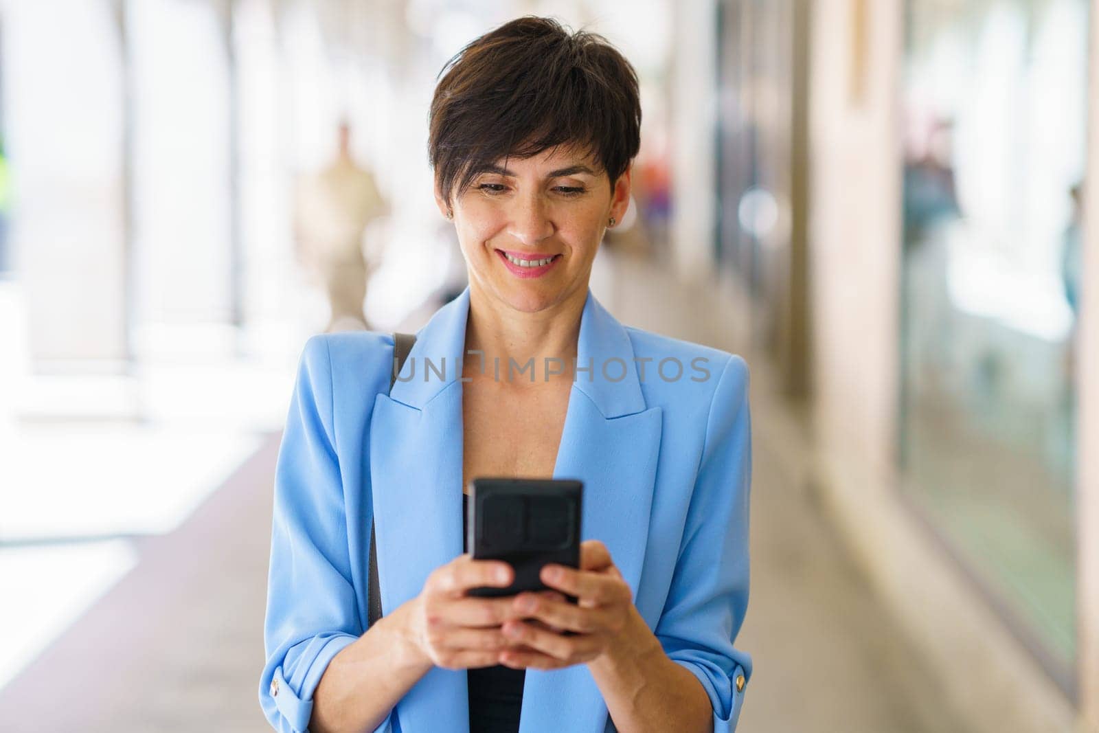 Cheerful middle aged female in blue jacket smiling and messaging on mobile phone while standing on sunny street during walk