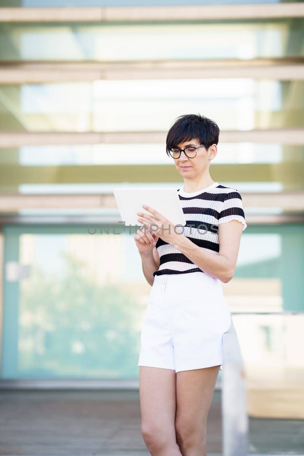 Side view of female in casual clothes standing near window and browsing tablet while working in modern office