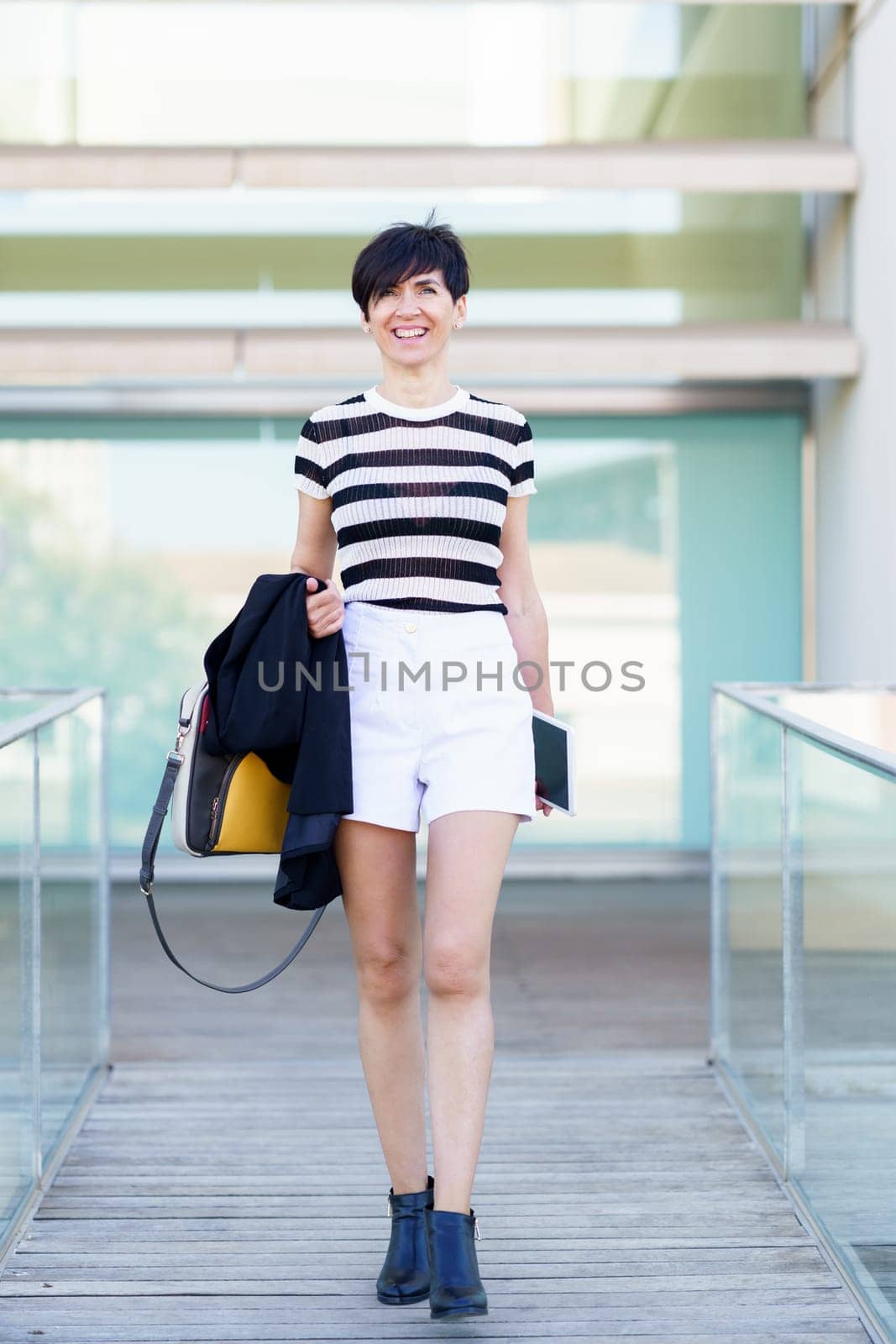 Full body of smiling female in striped shirt and shorts holding handbag while walking along corridor with tablet on blurred background