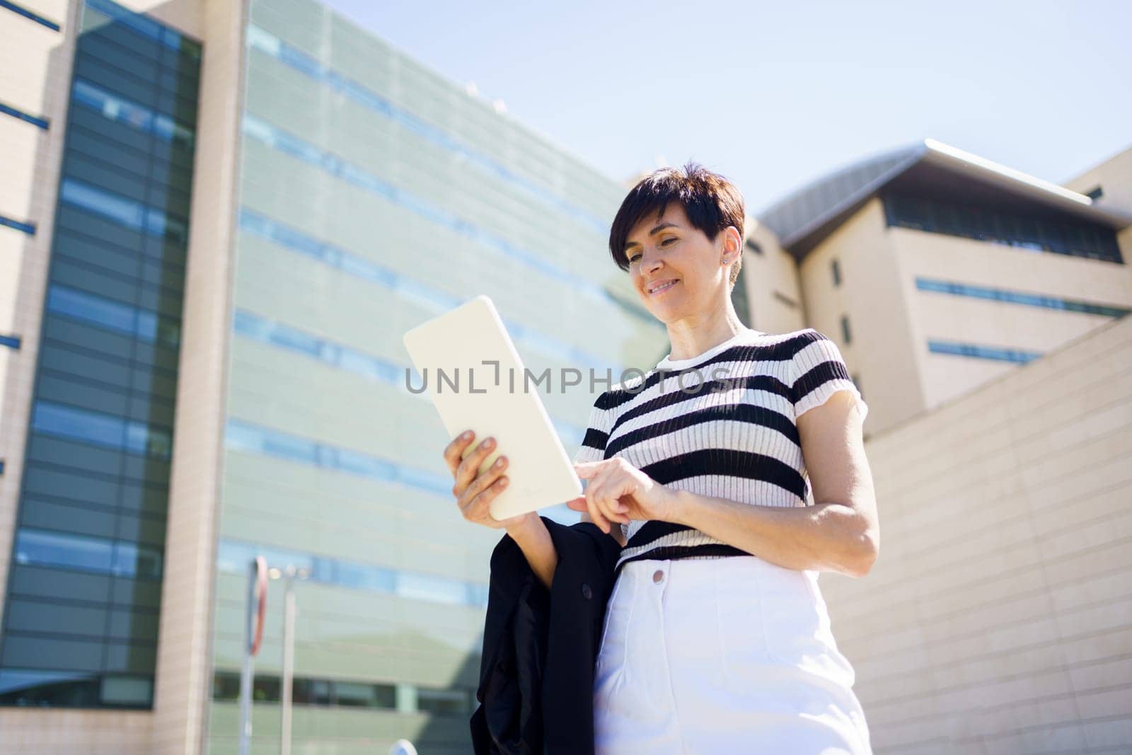 Low angle side view of female in casual clothes standing on street and reading notes in tablet while standing near modern building