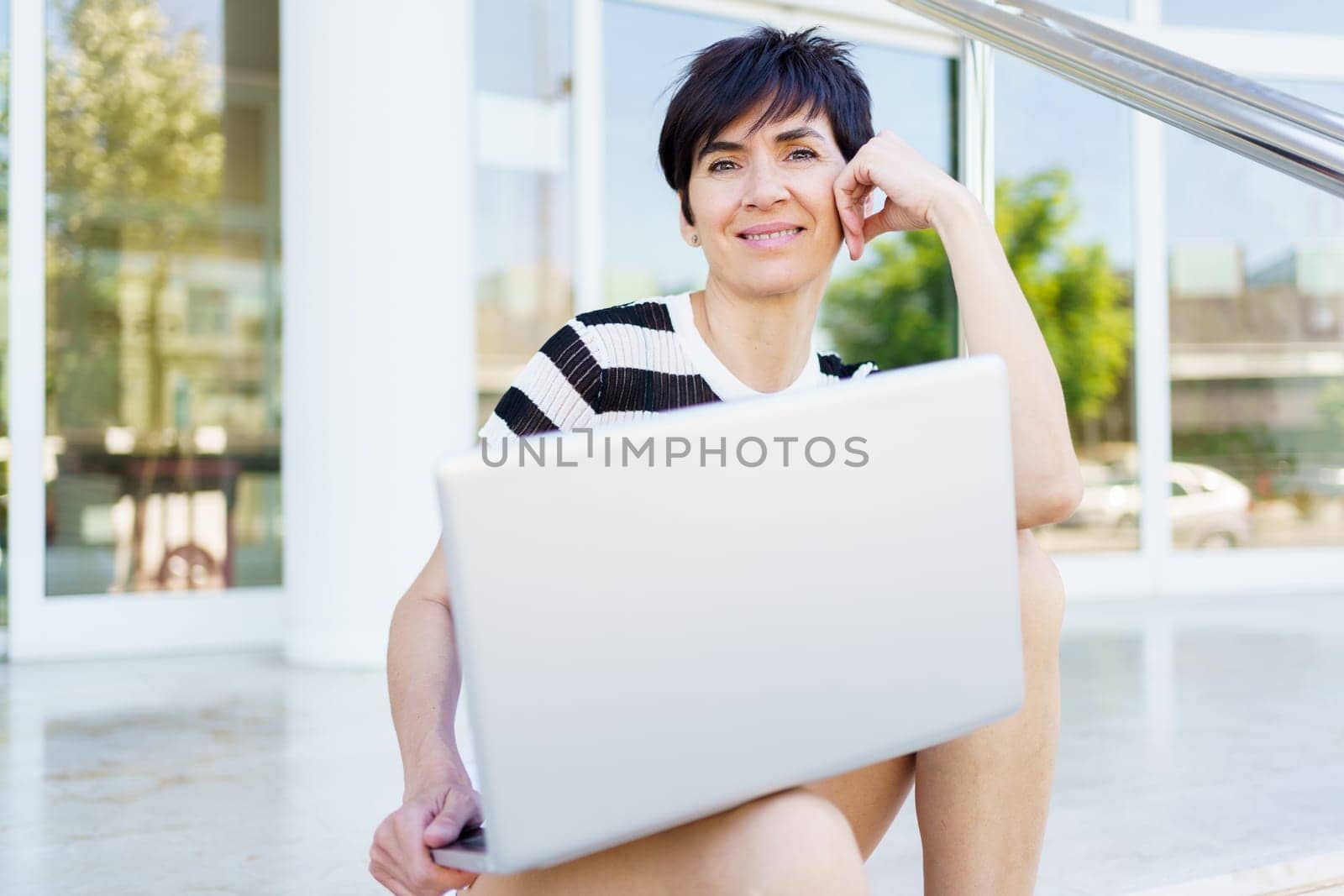 Cheerful adult female freelancer in striped t shirt looking at camera and smiling while sitting on street with laptop during remote work on project