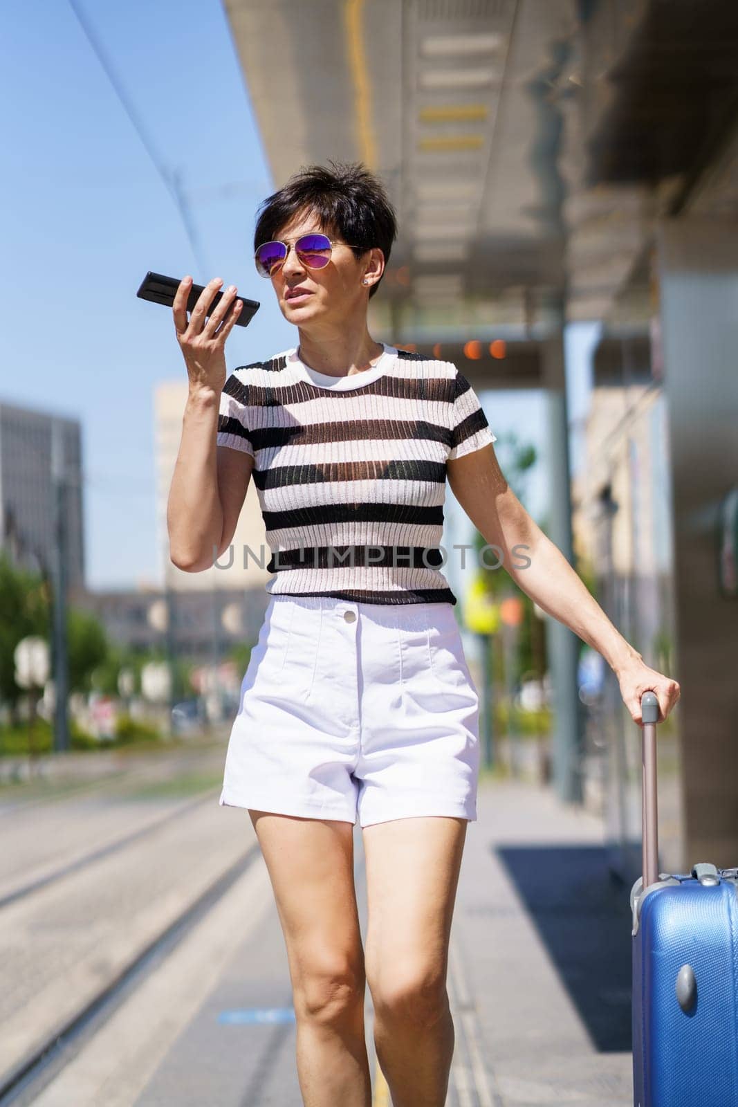 Positive young female in trendy striped t shirt with suitcase and sunglasses walking on city street by train station, while recording voice message on smartphone and looking away