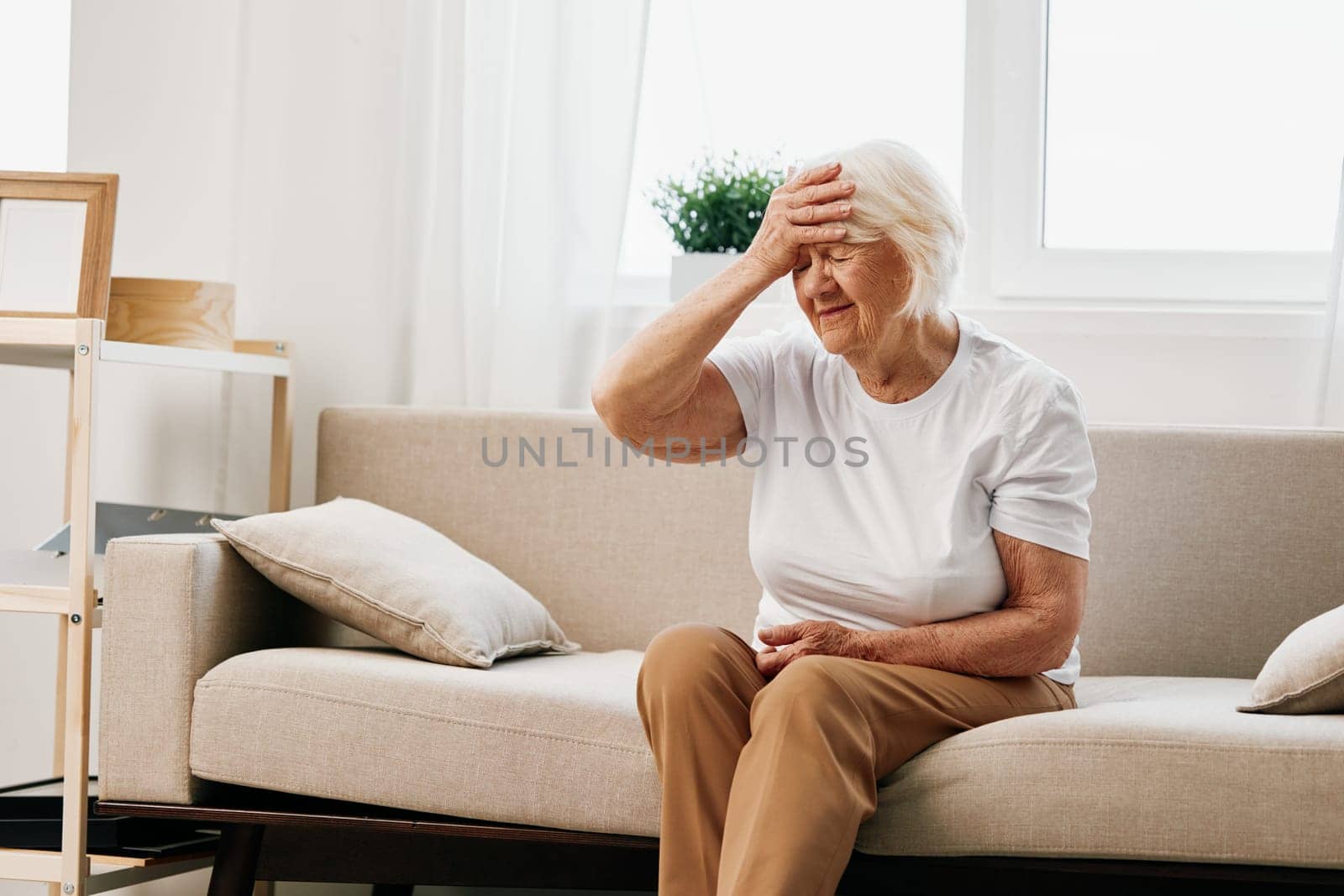 Elderly woman severe pain in the head sitting on the couch, health problems in old age, poor quality of life. Grandmother with gray hair holding her head, migraine and high blood pressure. High quality photo