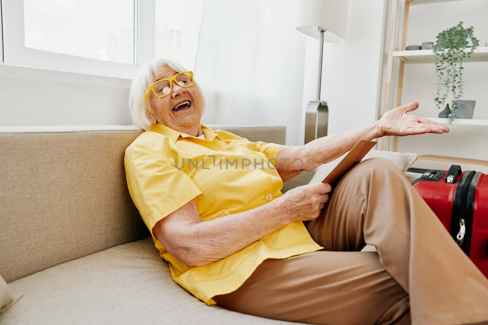 Happy senior woman with passport and travel ticket packed a red suitcase, vacation and health care. Smiling old woman joyfully sitting on the sofa before the trip raised her hands up in joy. High quality photo