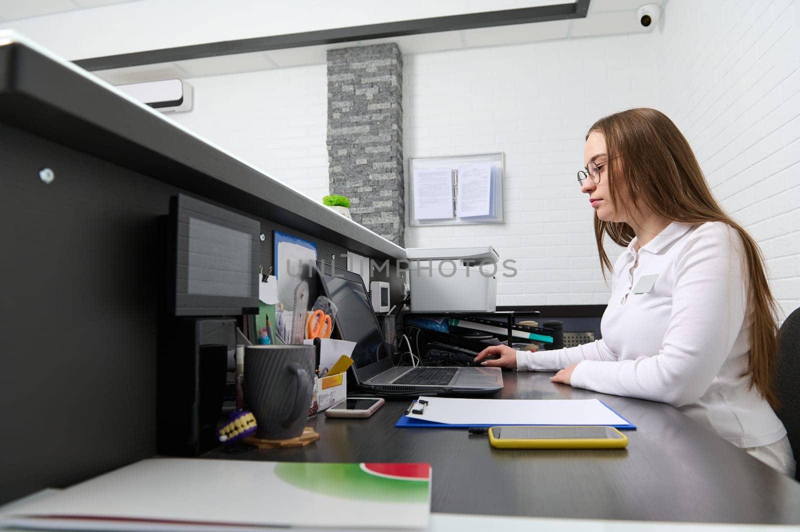 Portrait of a young woman in white uniform, receptionist administrator at reception desk, making appointments with the medical staff while sitting at laptop PC in modern dentistry clinic or spa salon