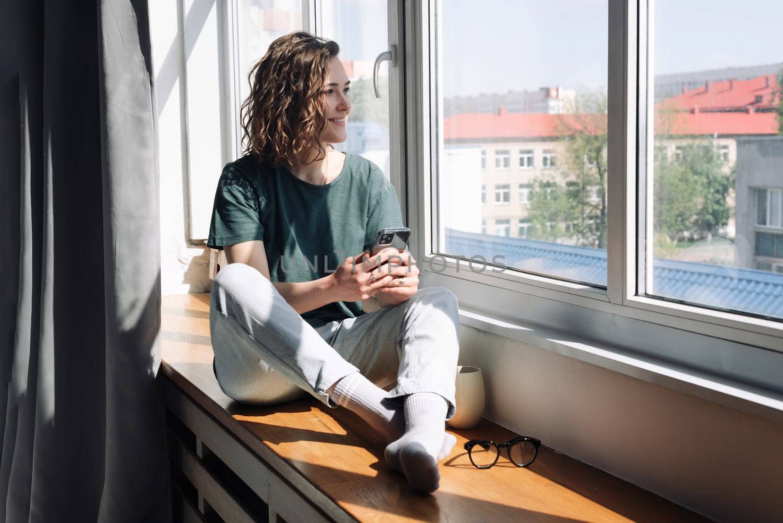 Digital Disconnect: Peaceful Young Woman Finds Serenity at Home, Gazing Out of Window, Balancing Technology and Relaxation in a Lockdown Era.
