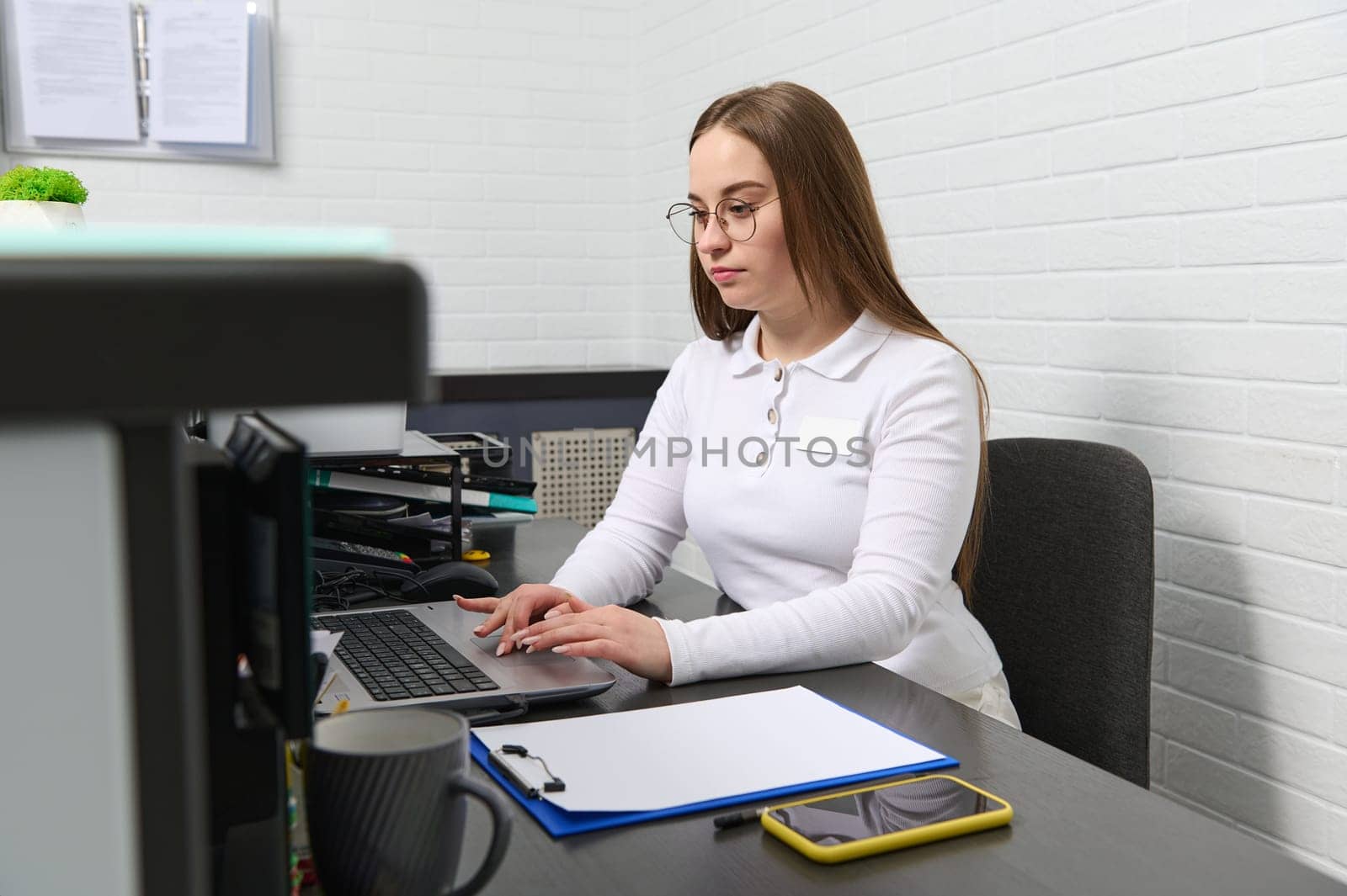 Receptionist works on laptop, sitting behind a reception desk in modern clinic, keeping appointments with medical staff by artgf