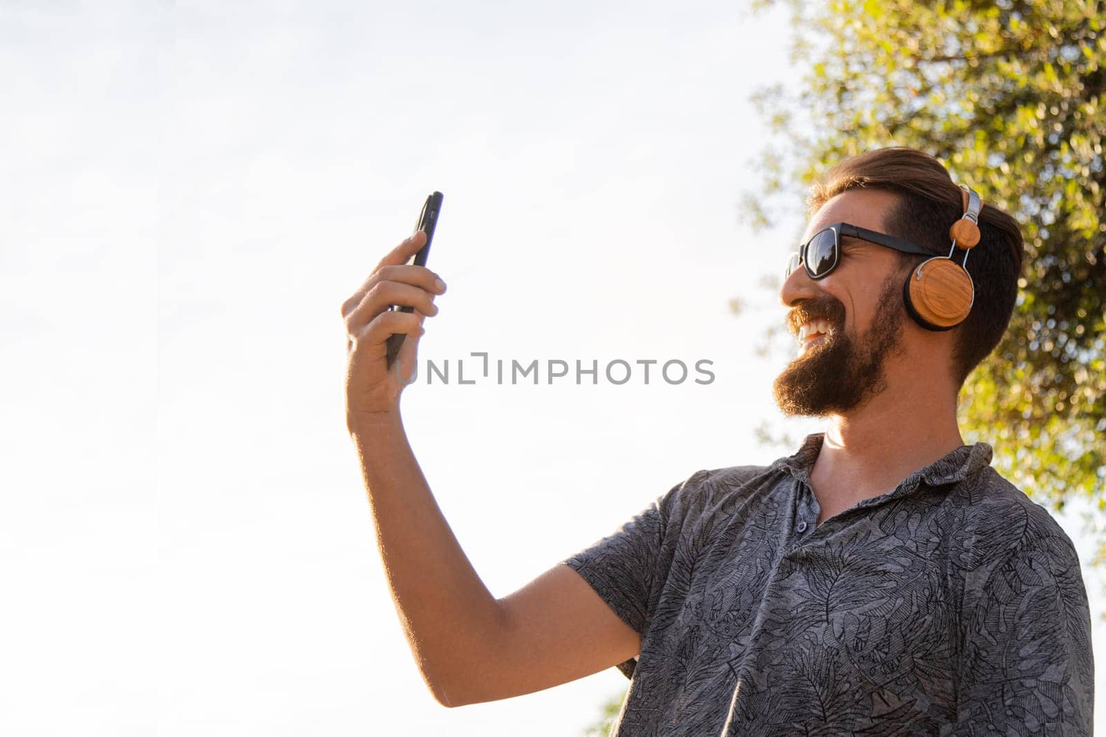 Young happy man taking selfie with mobile phone outdoors. Copy space. by Hoverstock