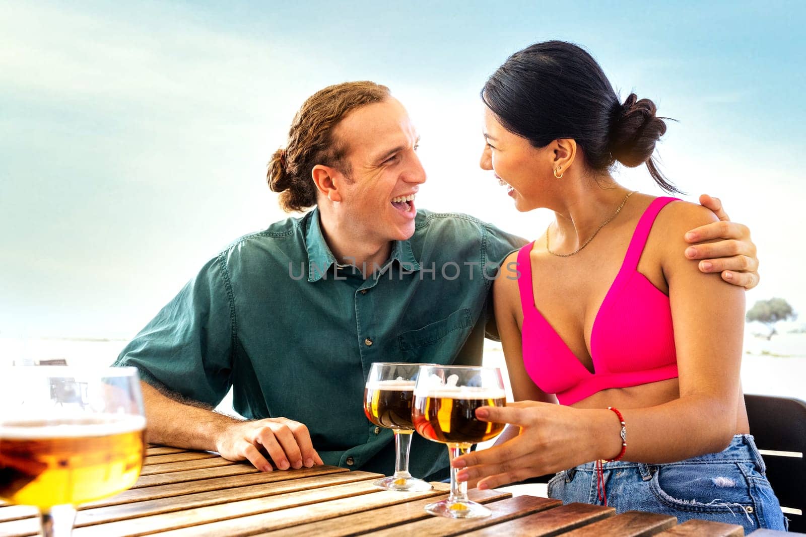 Happy multiracial couple on vacation having drinks together at a beach bar drinking beer. Lifestyle and summertime concept.