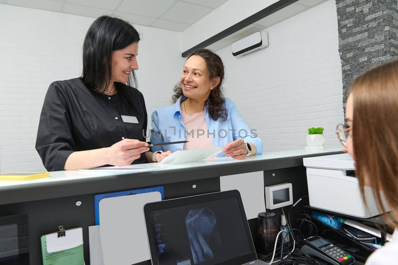 Female patient smiles, asking her doctor dentist informations filling in stomatological form, standing by reception desk by artgf