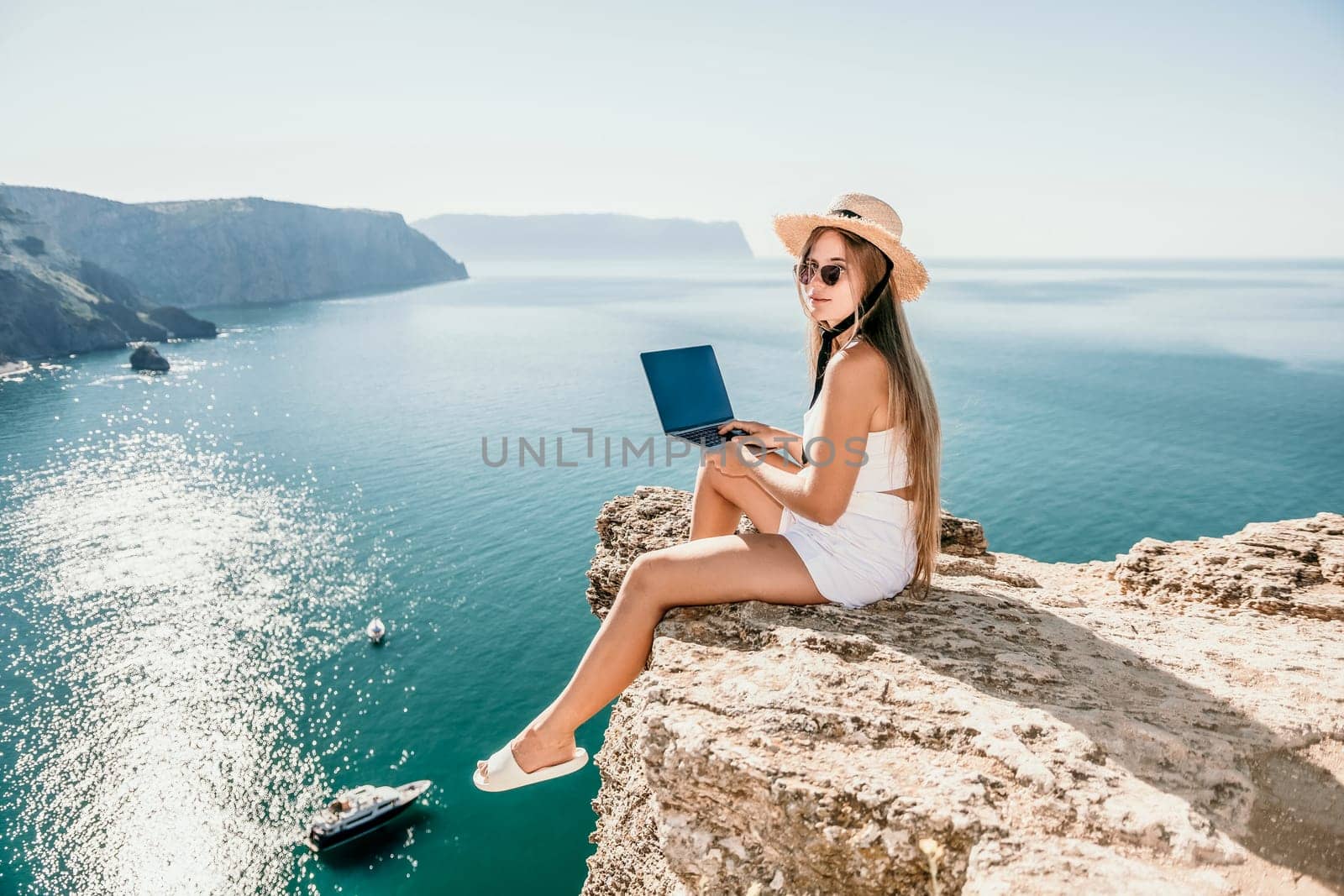 Successful business woman in yellow hat working on laptop by the sea. Pretty lady typing on computer at summer day outdoors. Freelance, travel and holidays concept.