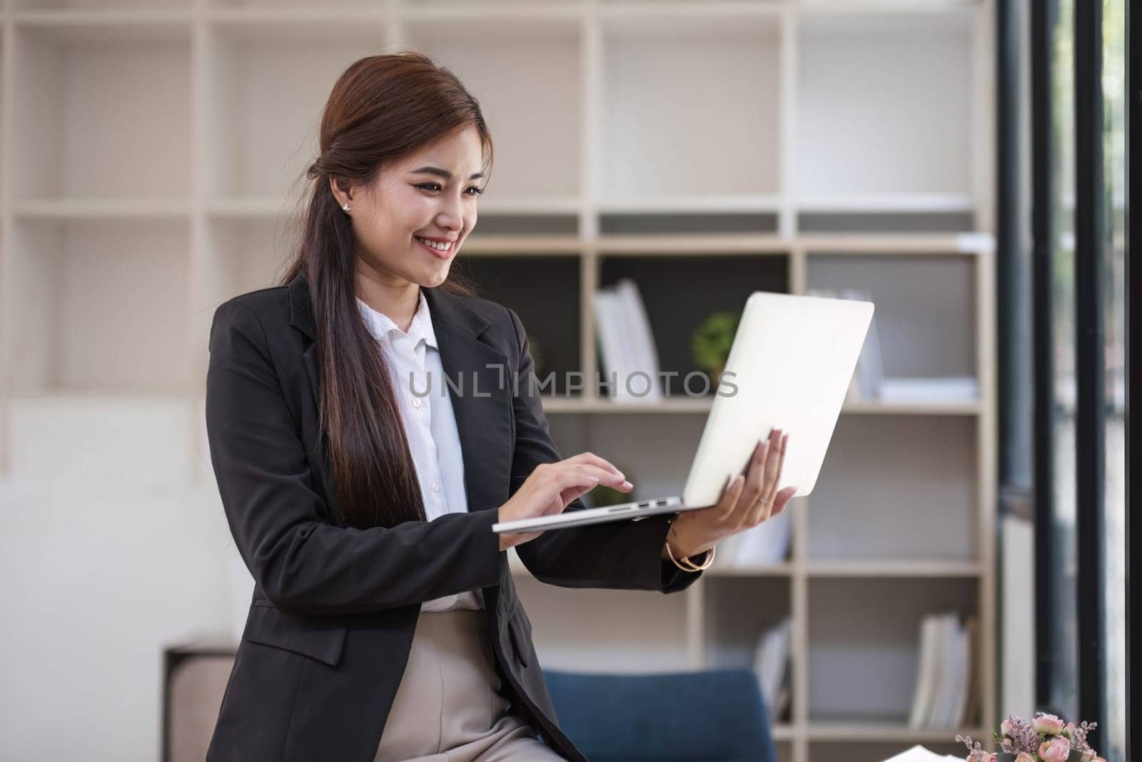 Portrait, Professional and confident millennial Asian businesswoman or female executive manager in formal suit and eyeglasses, standing, leaning on table, holding laptop and using laptop computer. by wichayada