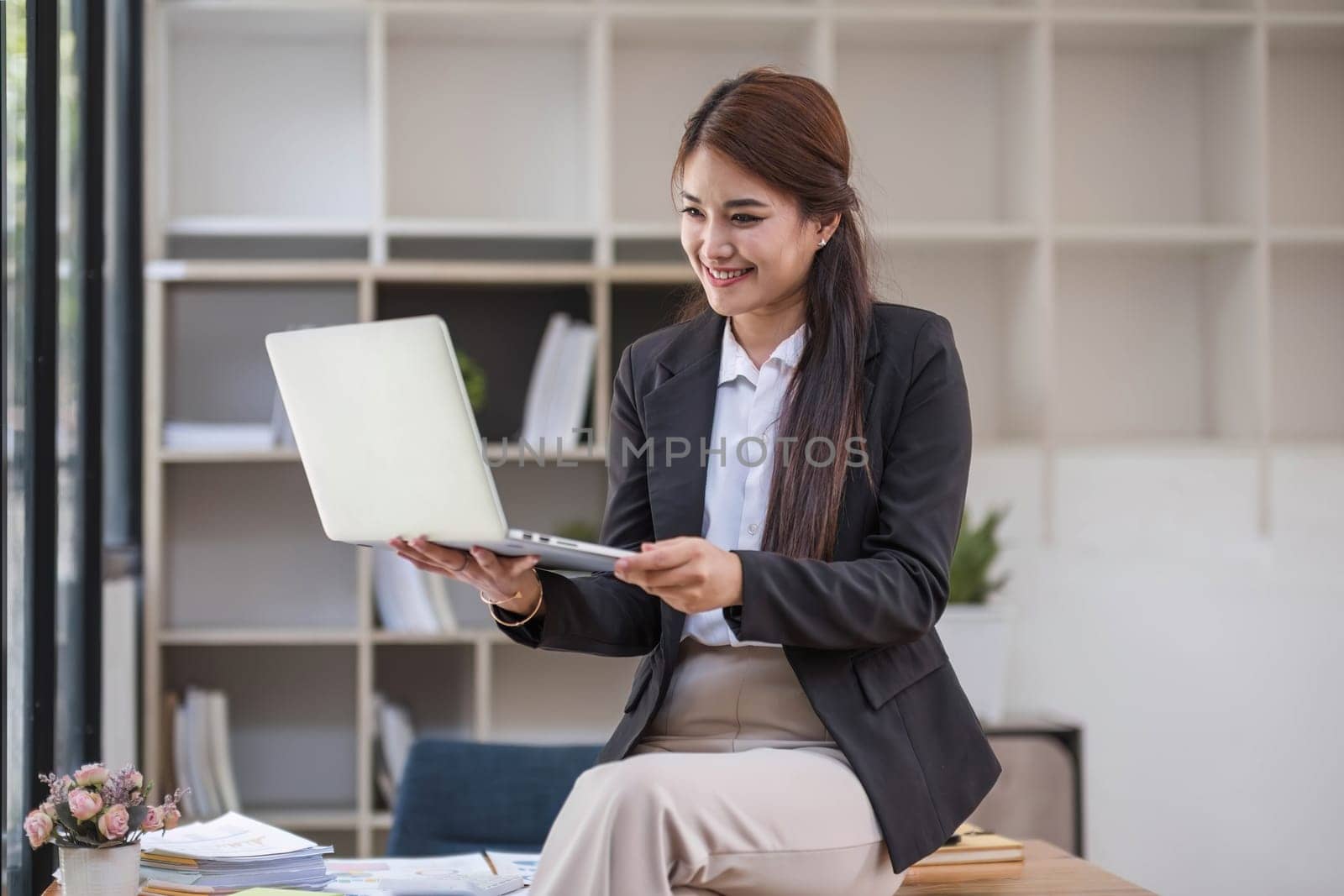 Portrait, Professional and confident millennial Asian businesswoman or female executive manager in formal suit and eyeglasses, standing, leaning on table, holding laptop and using laptop computer...