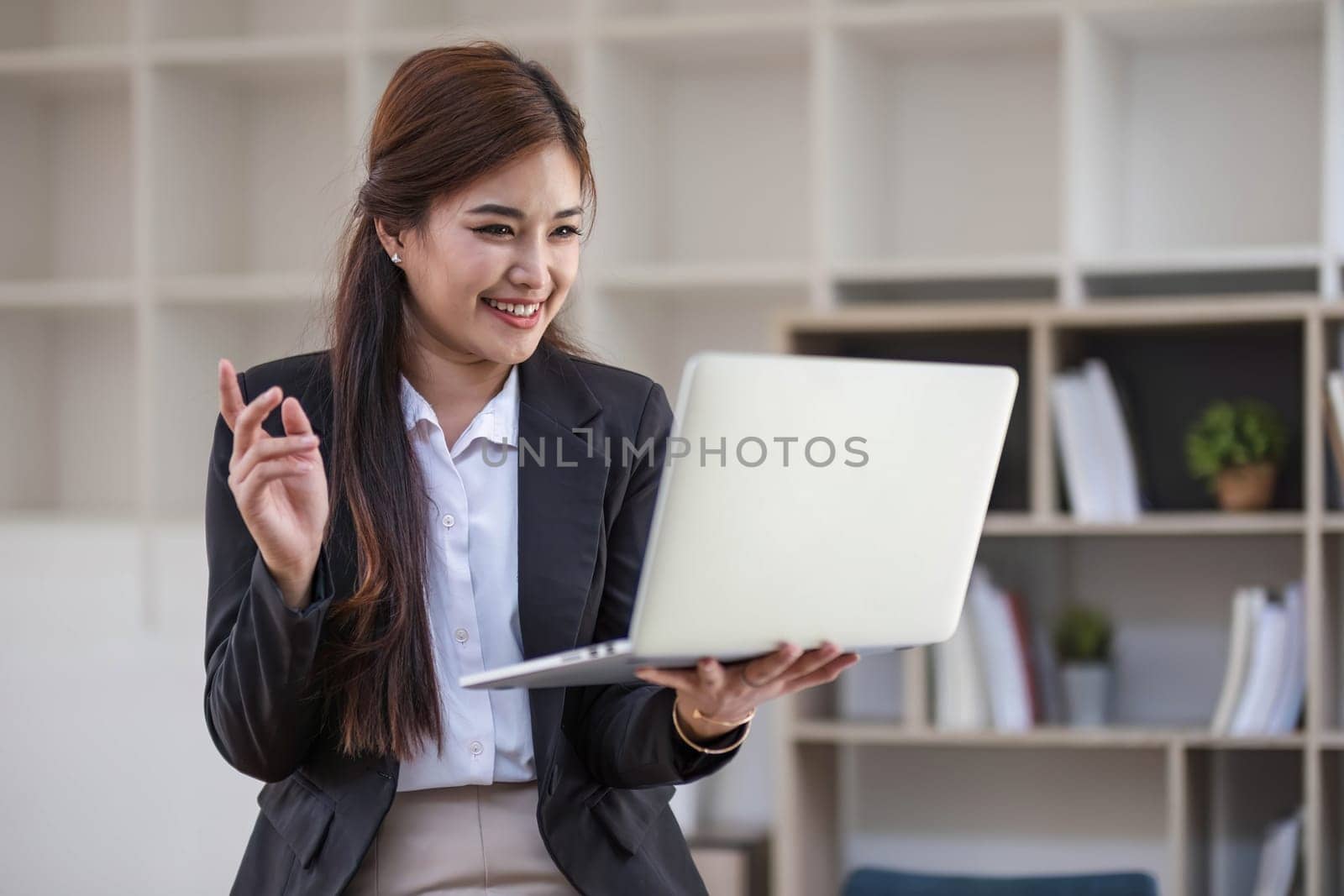 Portrait, Professional and confident millennial Asian businesswoman or female executive manager in formal suit and eyeglasses, standing, leaning on table, holding laptop and using laptop computer...