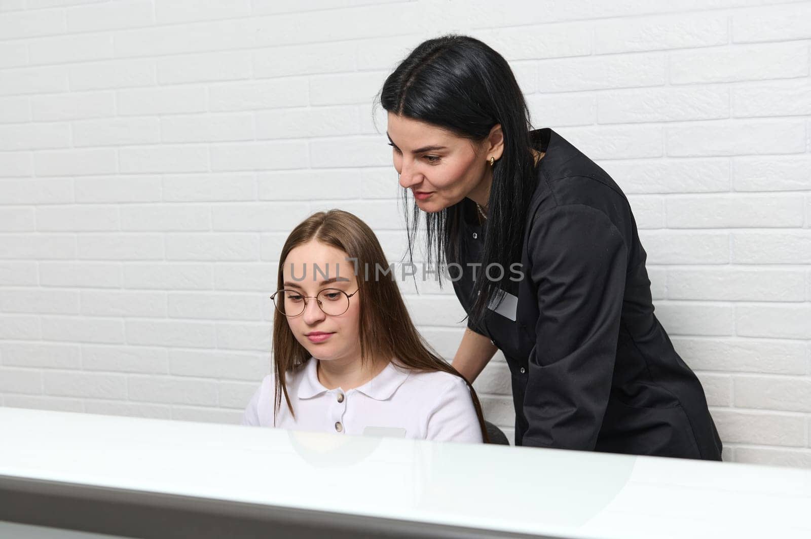 Young confident beautiful female doctor scheduling appointment, standing near receptionist by reception desk in clinic by artgf