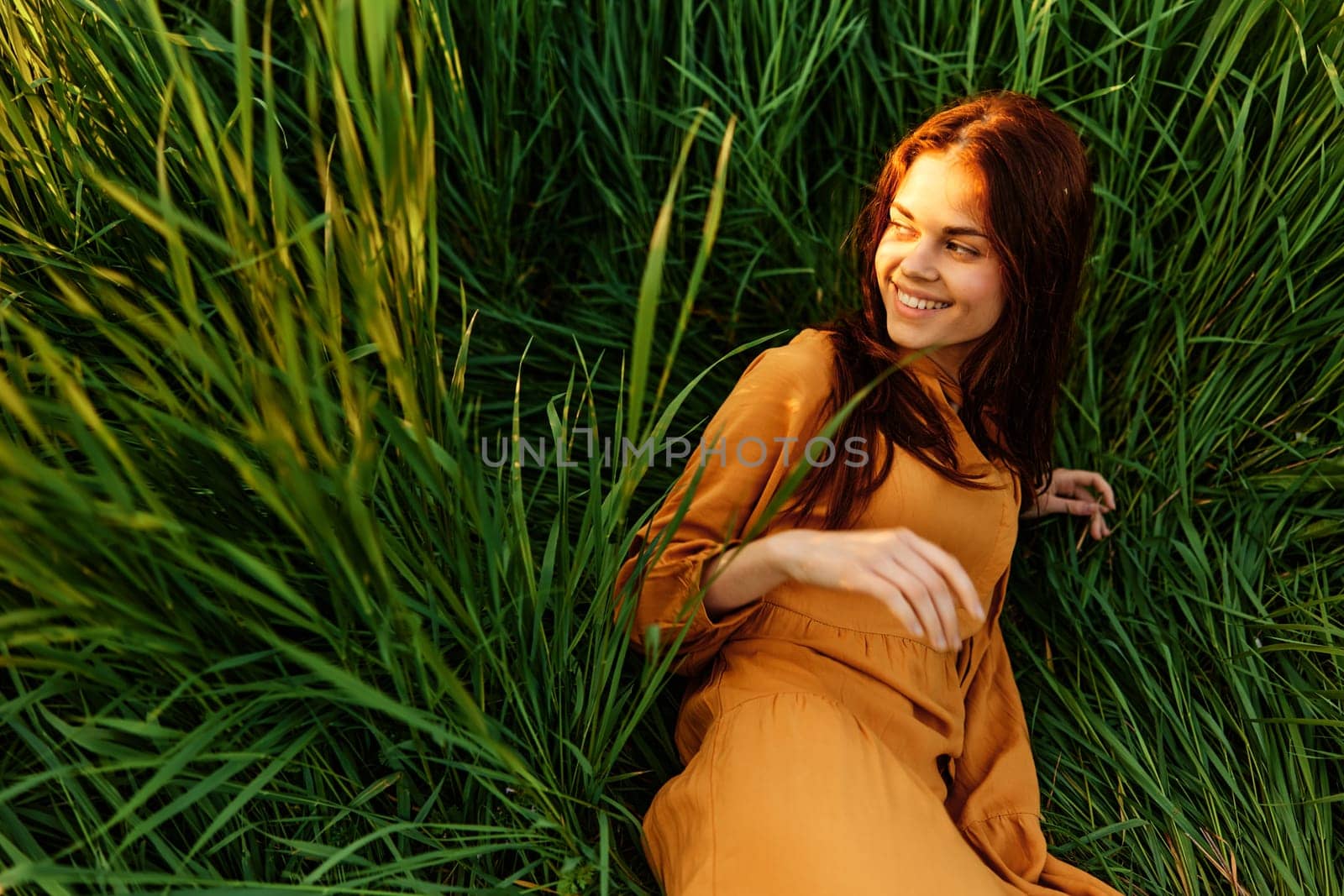 a happy woman lies in the tall green grass in a long orange dress and looks away smiling pleasantly. High quality photo