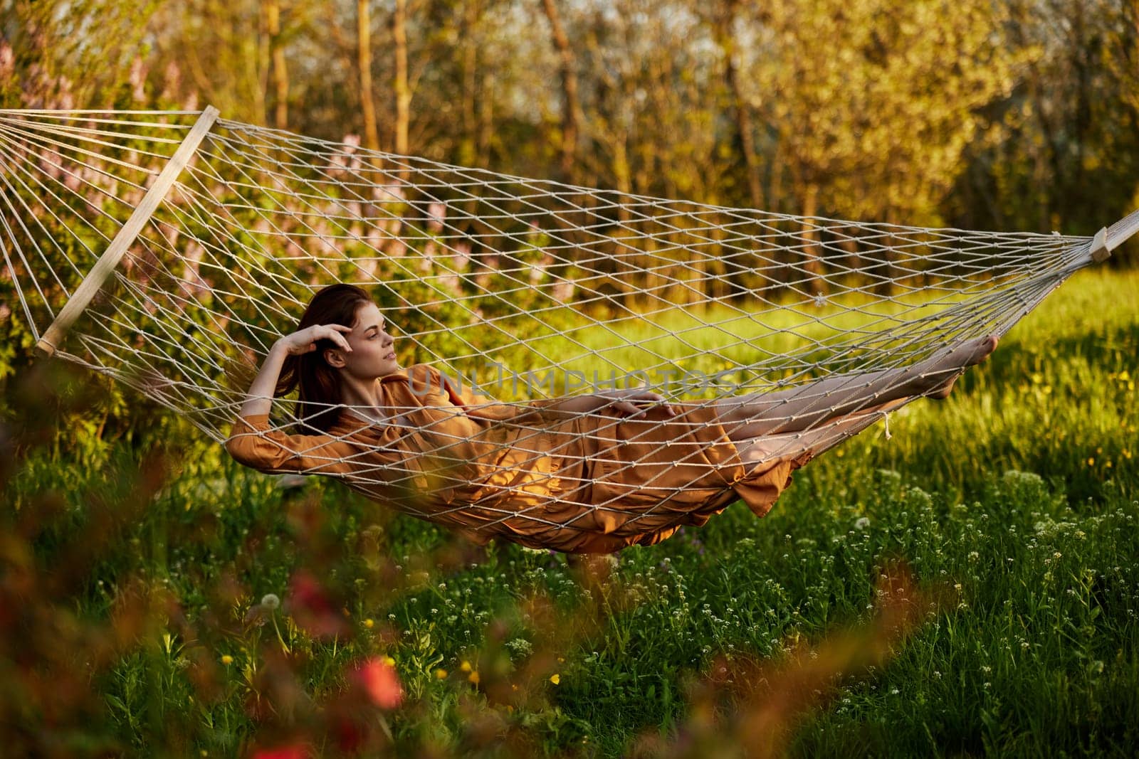 a beautiful woman is resting in nature lying in a mesh hammock in a long orange dress looking to the side, propping her head with her hand. Horizontal photo on the theme of recreation. High quality photo