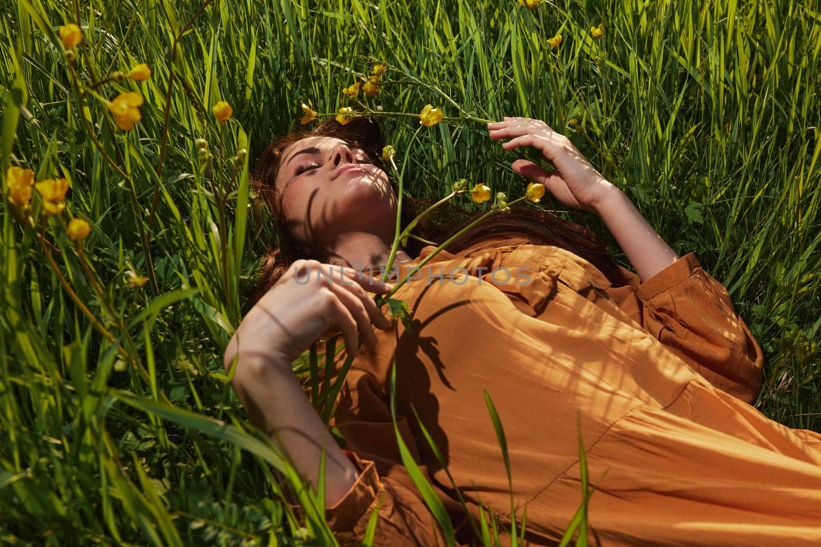 a calm woman with long red hair lies in a green field with yellow flowers, in an orange dress with her eyes closed, with a pleasant smile on her face, enjoying peace and recuperating, illuminated by the setting sun. High quality photo