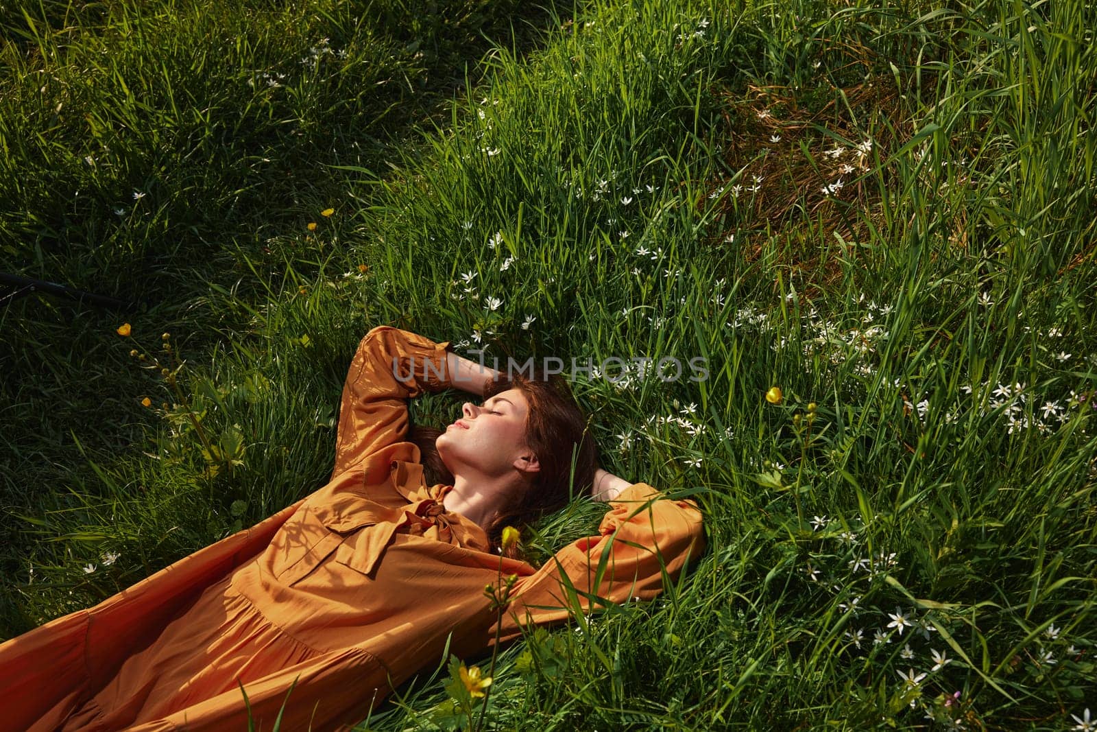 a happy, relaxed woman, resting lying in the green grass, in a long orange dress, with her eyes closed and a pleasant smile on her face, enjoying harmony with nature and recuperating by Vichizh