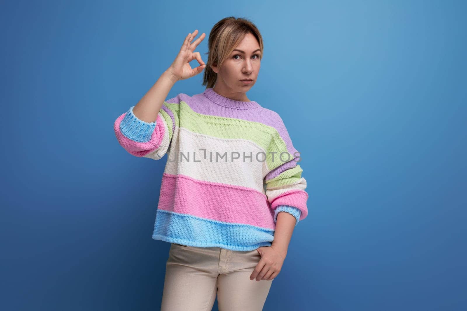 serious blond young woman in a striped sweater shows with her hand a class in on a blue background.